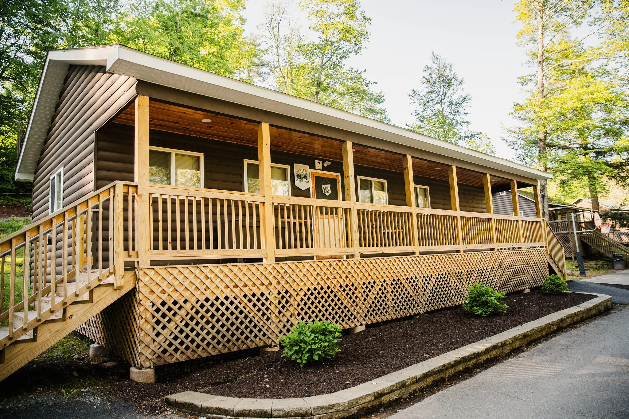 Front view of a single-story cabin with a wooden porch, surrounded by greenery and trees.