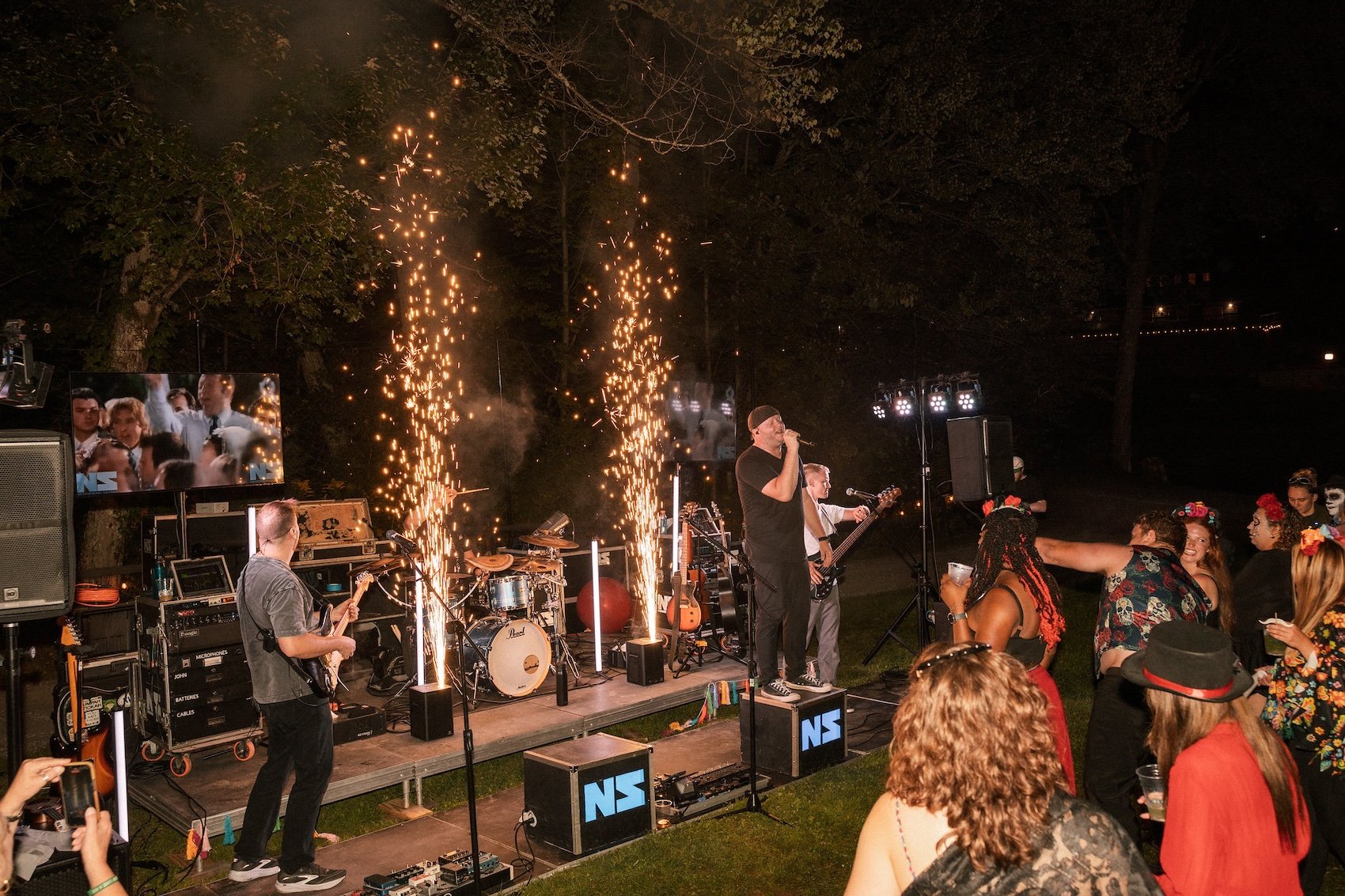 A live outdoor musical performance at night with a band on stage, pyrotechnics, and an audience enjoying the show. The singer is wearing black and singing into a microphone, surrounded by band members playing guitar and drums. The scene is lit with s