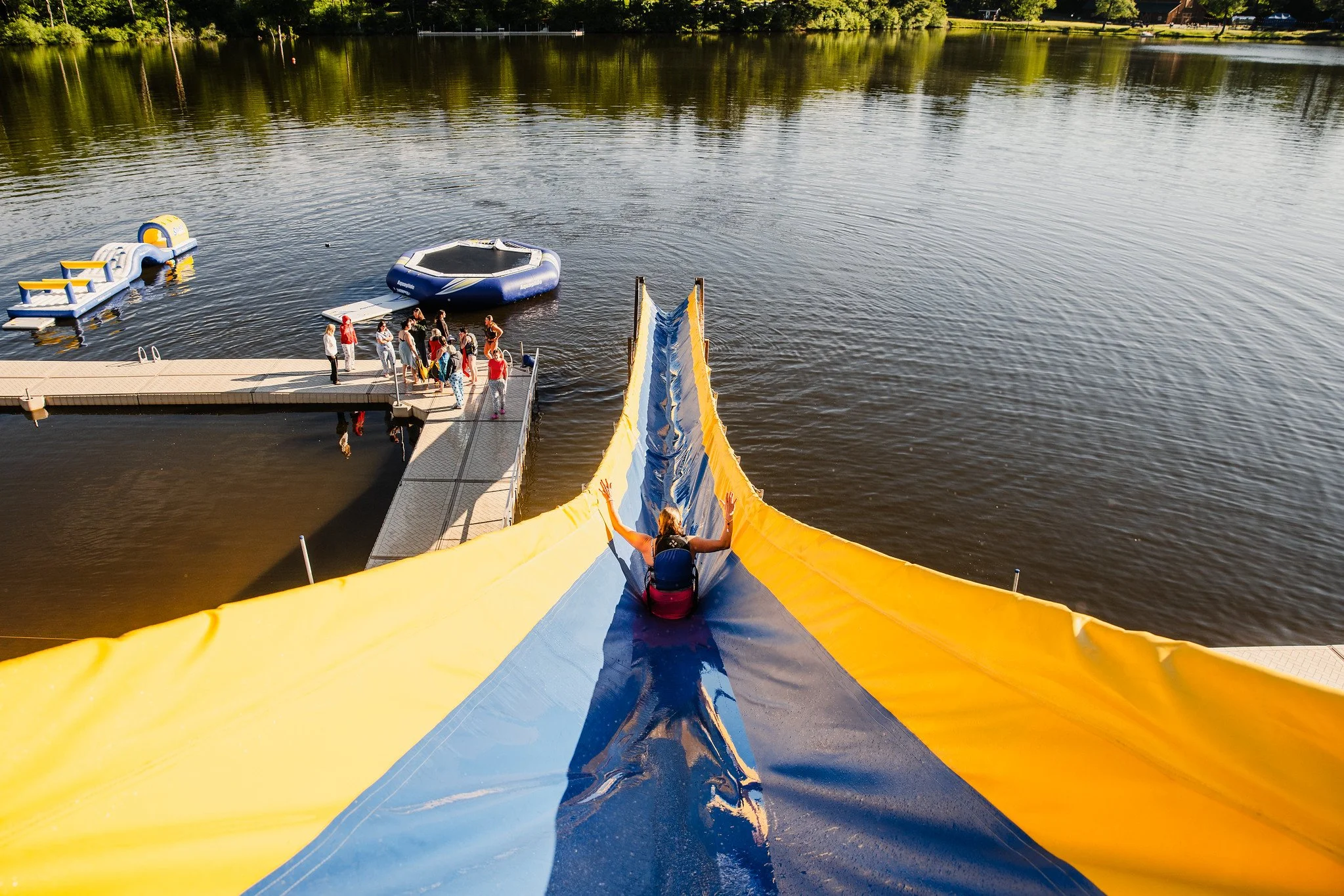 People gathered at a dock preparing for a ride down a water slide on a lake, with inflatable devices and a slide in the foreground.