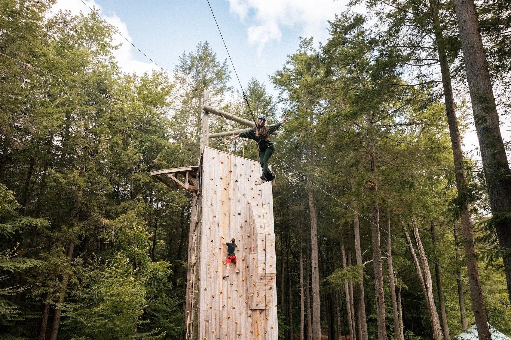Person on a zip line above a tall wooden climbing wall in a forested area, with a person climbing the wall and another person standing near the top.