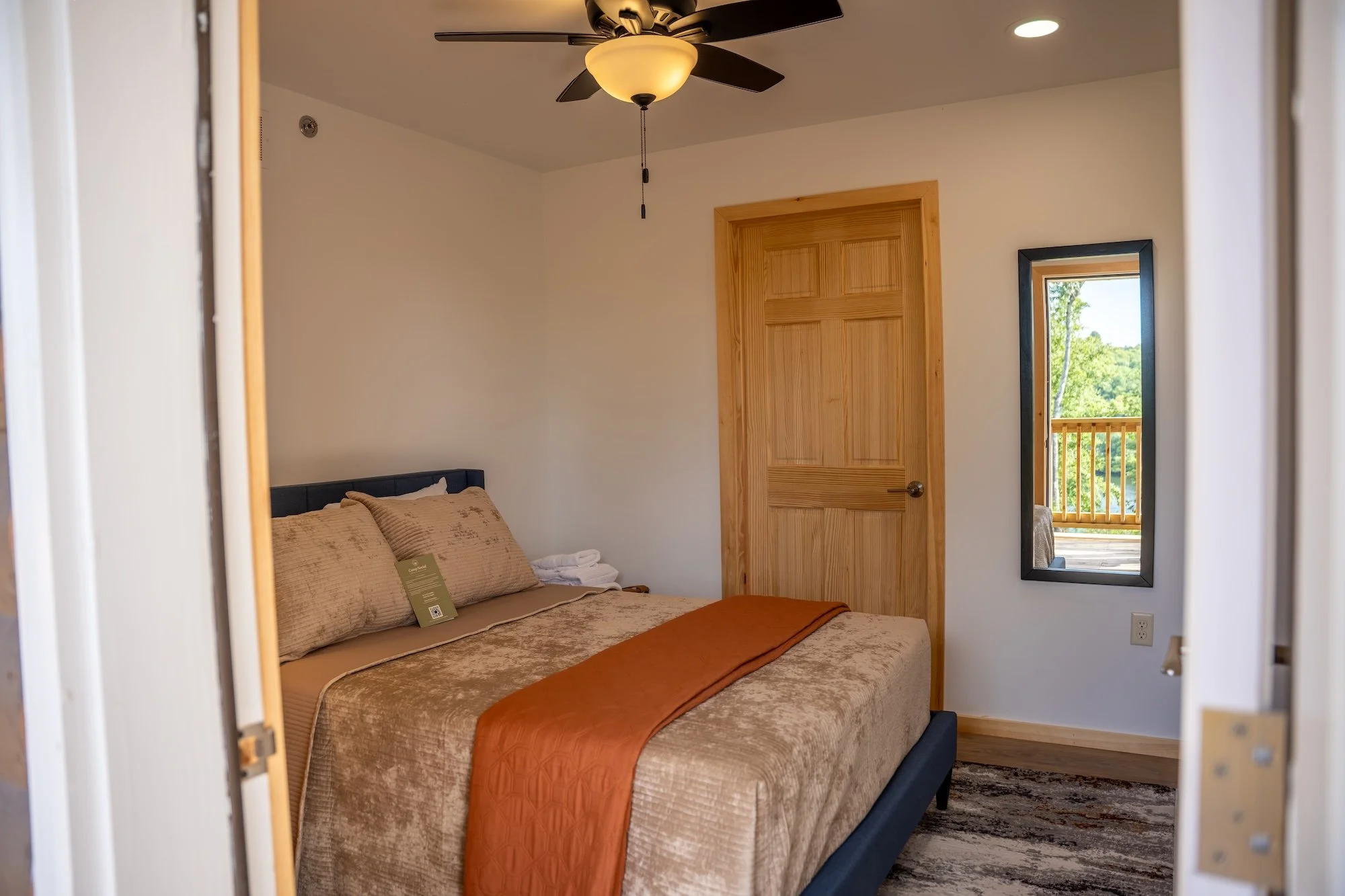 Wood-paneled bedroom with a bed, blue bedspread, gray pillows, a window with light blue curtains, a white dresser, a black mini fridge, a patterned armchair with a dark pillow, a floor lamp, and a small white ceiling vent.