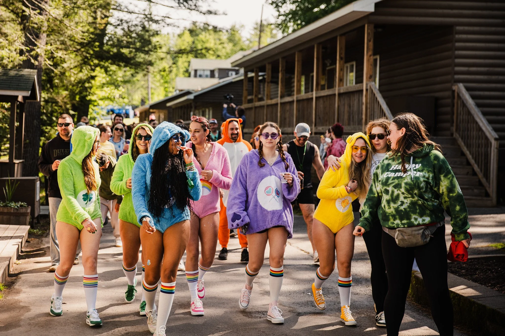 Group of people, mostly young women, walking outdoors wearing colorful, cozy hoodies, shorts, and knee-high socks with rainbow stripes, enjoying a sunny day at a camp or festival.