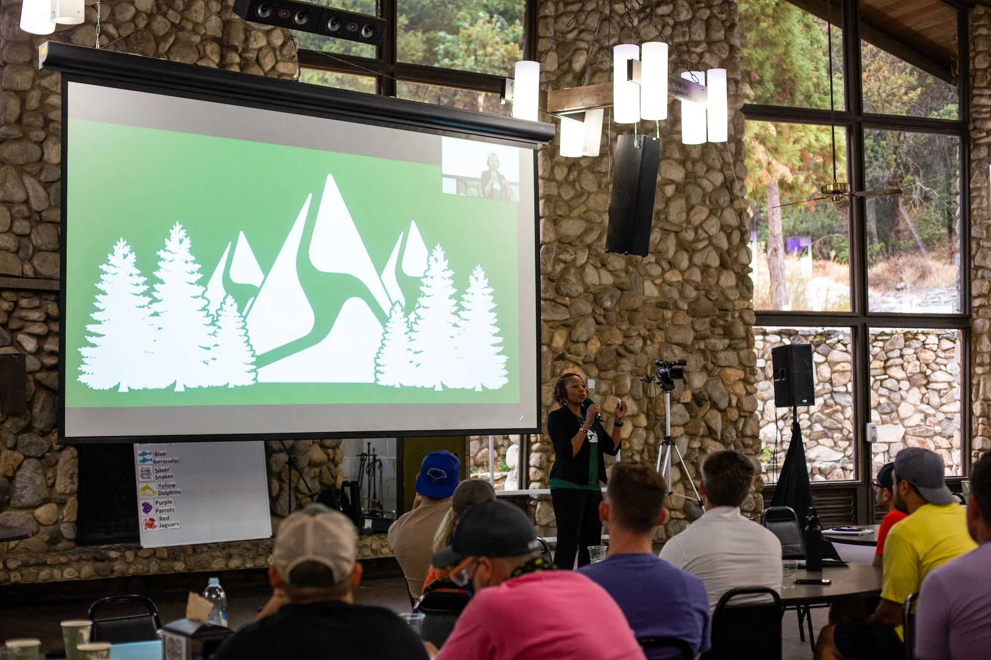 A woman giving a presentation to a group of people in a room with stone walls and large windows, featuring a large screen displaying a mountain and trees graphic.