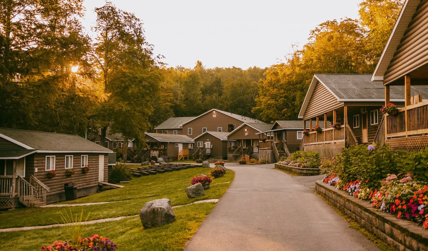 A scenic view of a small community with wooden houses on a peaceful street at sunset, surrounded by autumn trees and blooming flower beds.