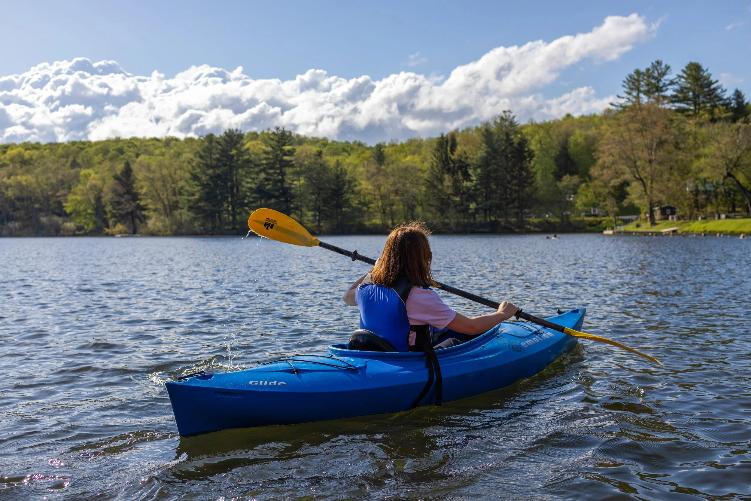 A person kayaking on a lake surrounded by green trees under a partly cloudy sky.