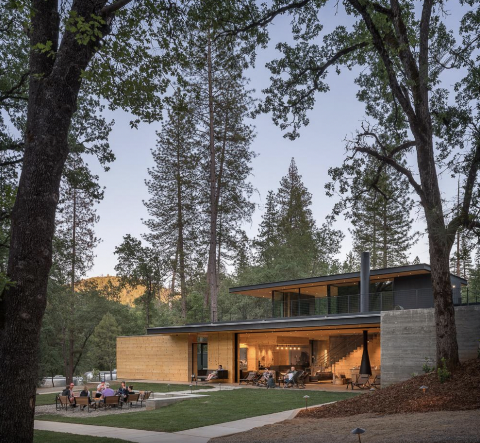 Modern house with large outdoor patio in a forested area, people seated and socializing on the patio during evening