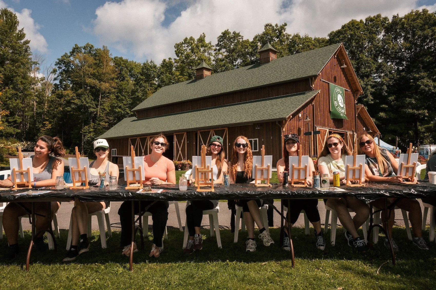 A group of women sitting at an outdoor table with mini easels, smiling, in front of a rustic barn with green trees in the background.