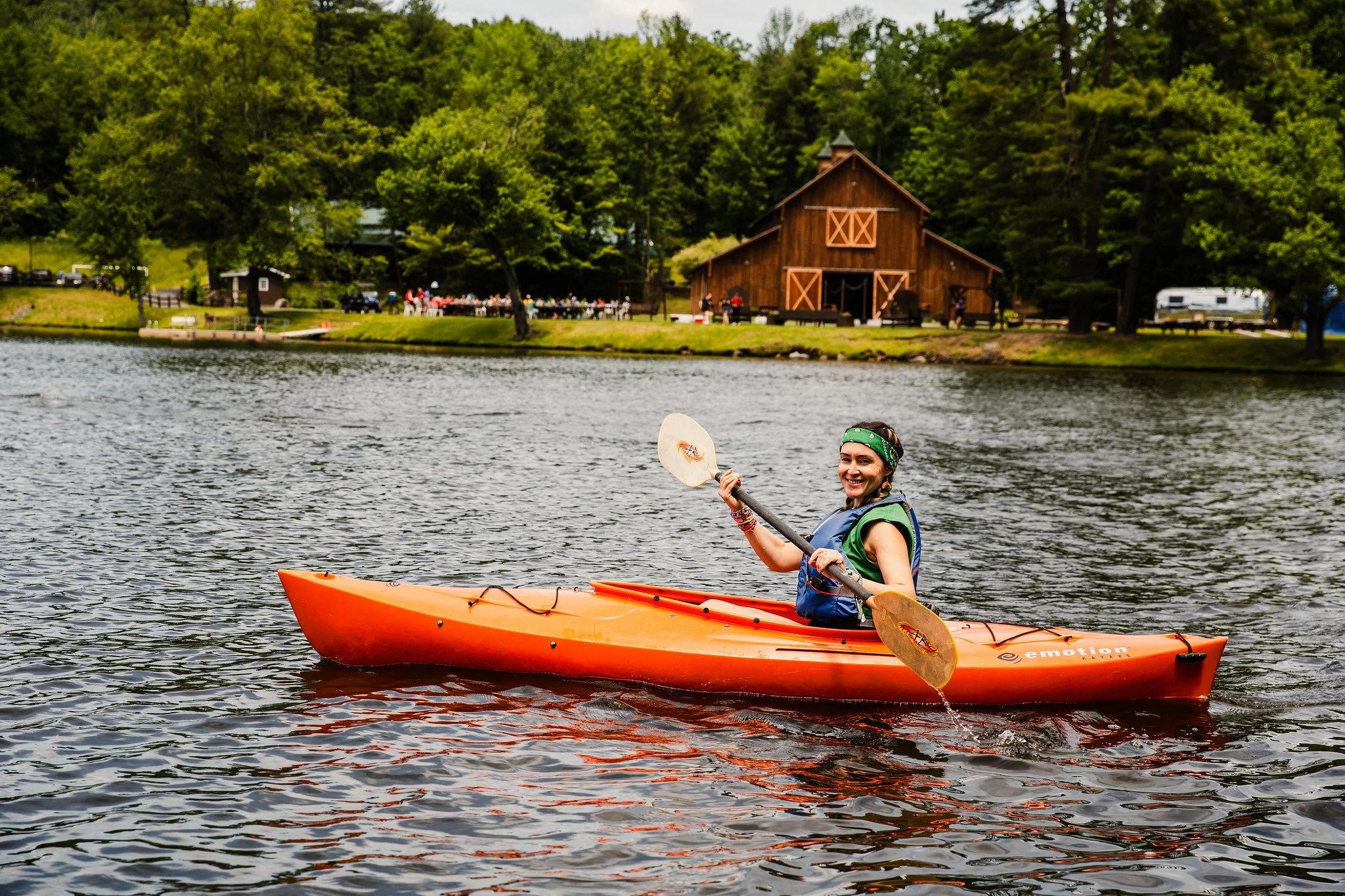 Smiling woman in a blue life jacket kayaking on a lake, surrounded by green trees and a rustic wooden barn in the background.