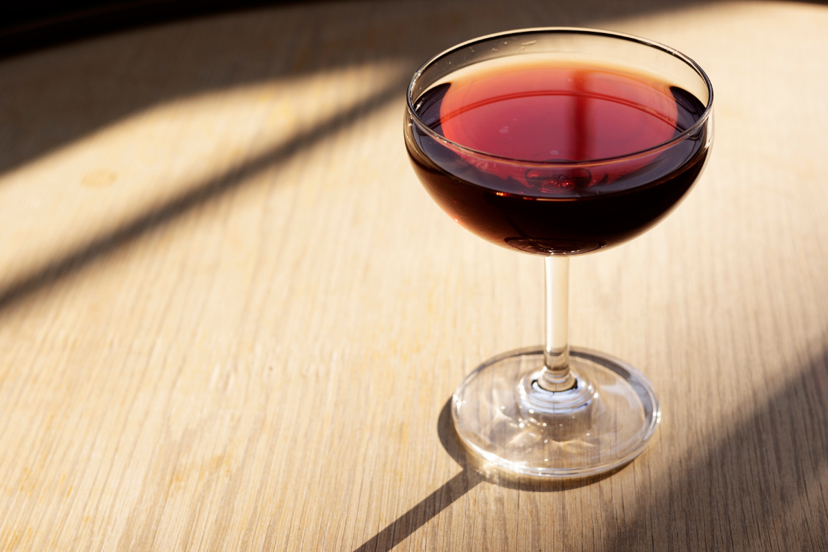 A dark red cocktail in a coupe glass sits atop a light wooden table, shadows cast from the top right of the image