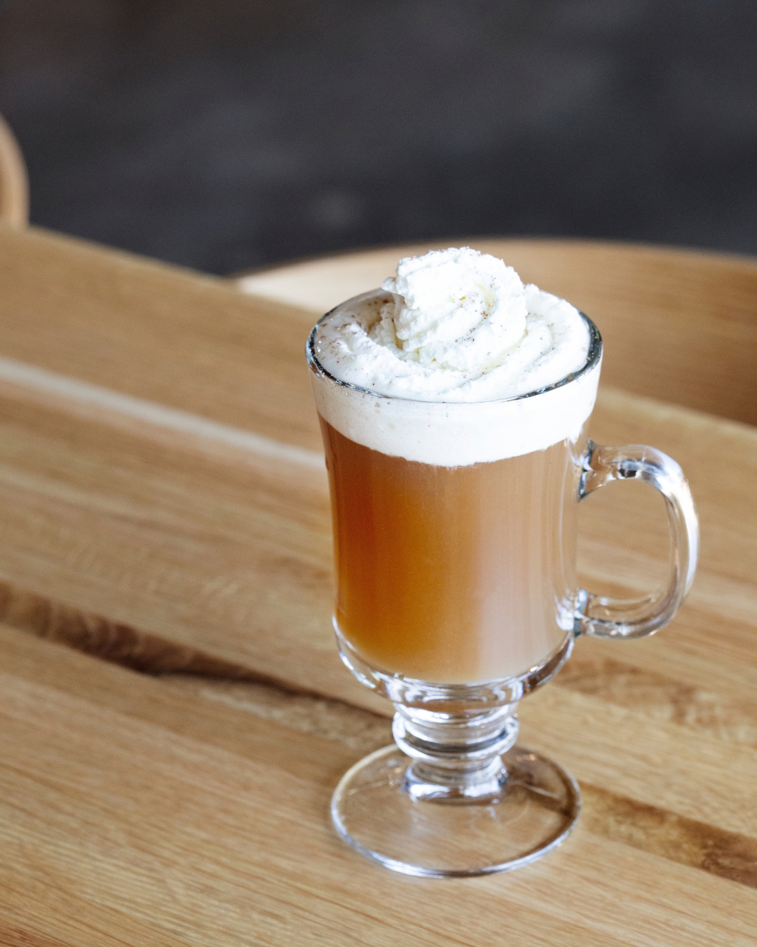 A warmed cider cocktail with a dollop of whipped cream in an Irish coffee-style mug sits atop a wooden table