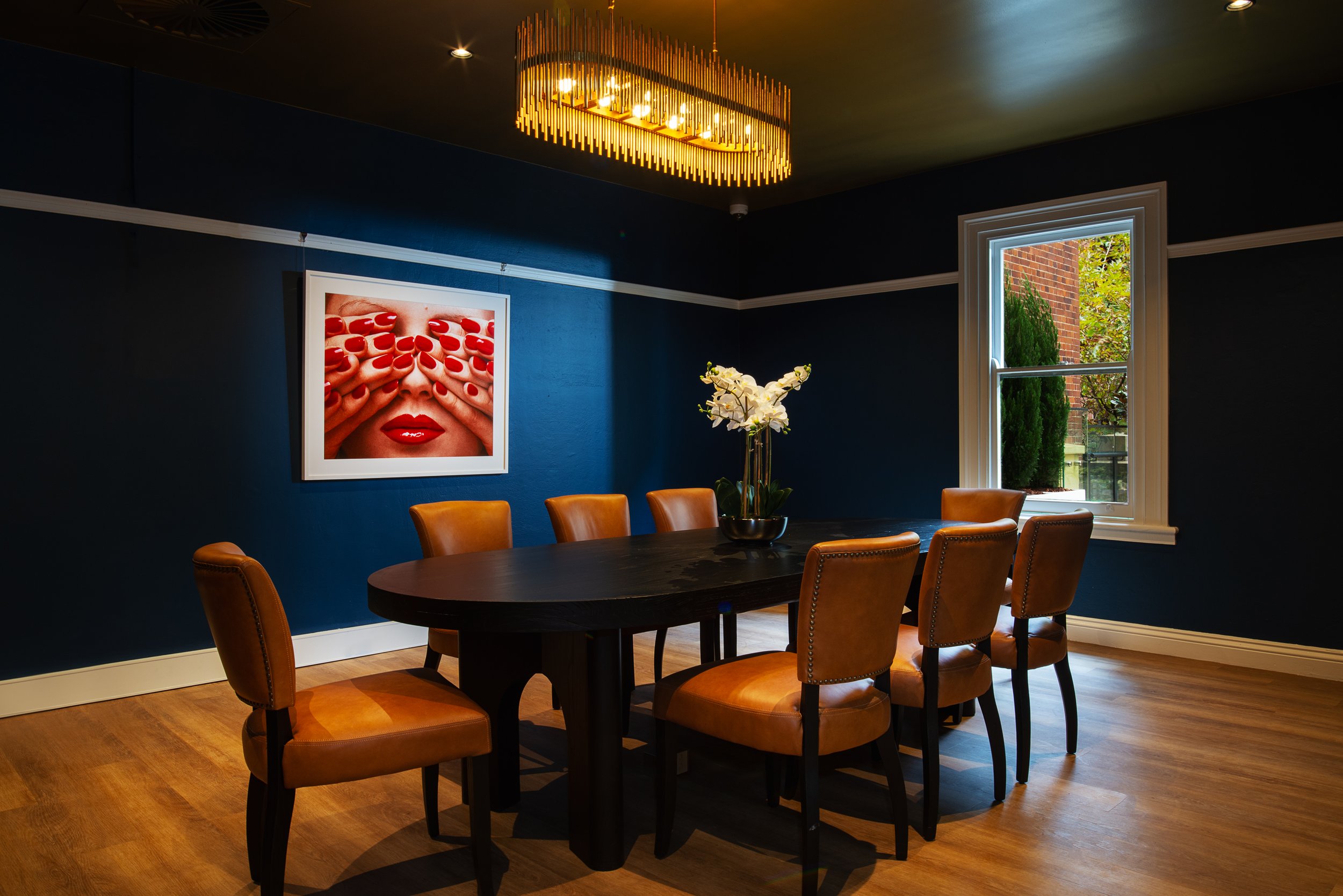 A dining room with dark blue walls, a wooden floor, and a window showing trees outside. There is a black oval table with seven tan leather chairs, a vase with white flowers, a picture on the wall of red nails covering eyes and lips, and a gold chande