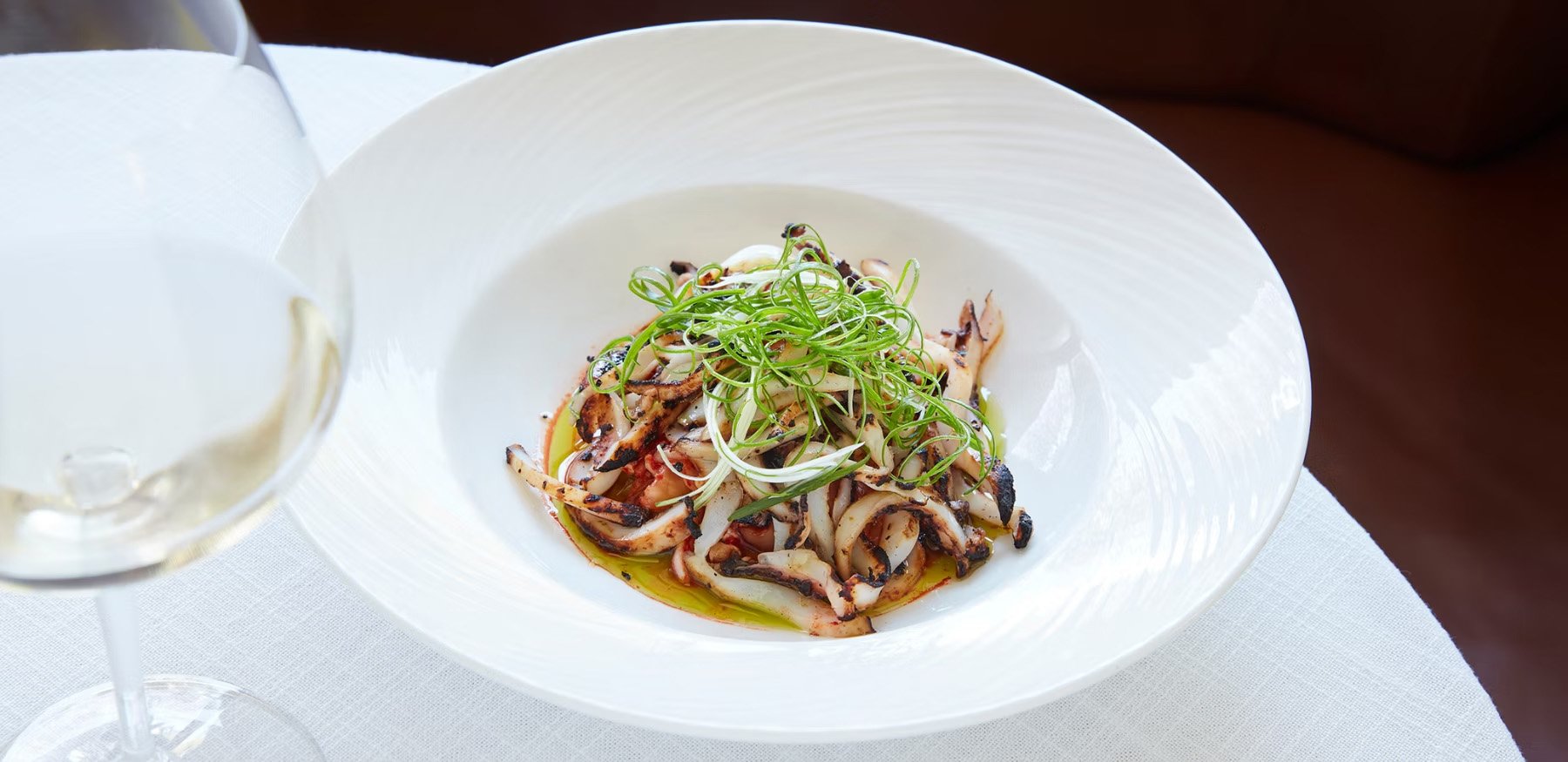 A white bowl with grilled calamari topped with chopped green herbs and garnished with thinly sliced green onions, sitting on a white tablecloth. A glass of white wine is partially visible on the left.