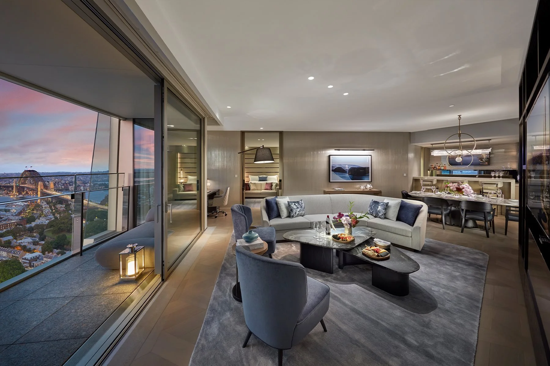 Modern living room with large windows overlooking a cityscape at sunset, featuring a sectional sofa, coffee table, dining area with chairs, and artwork on the walls.
