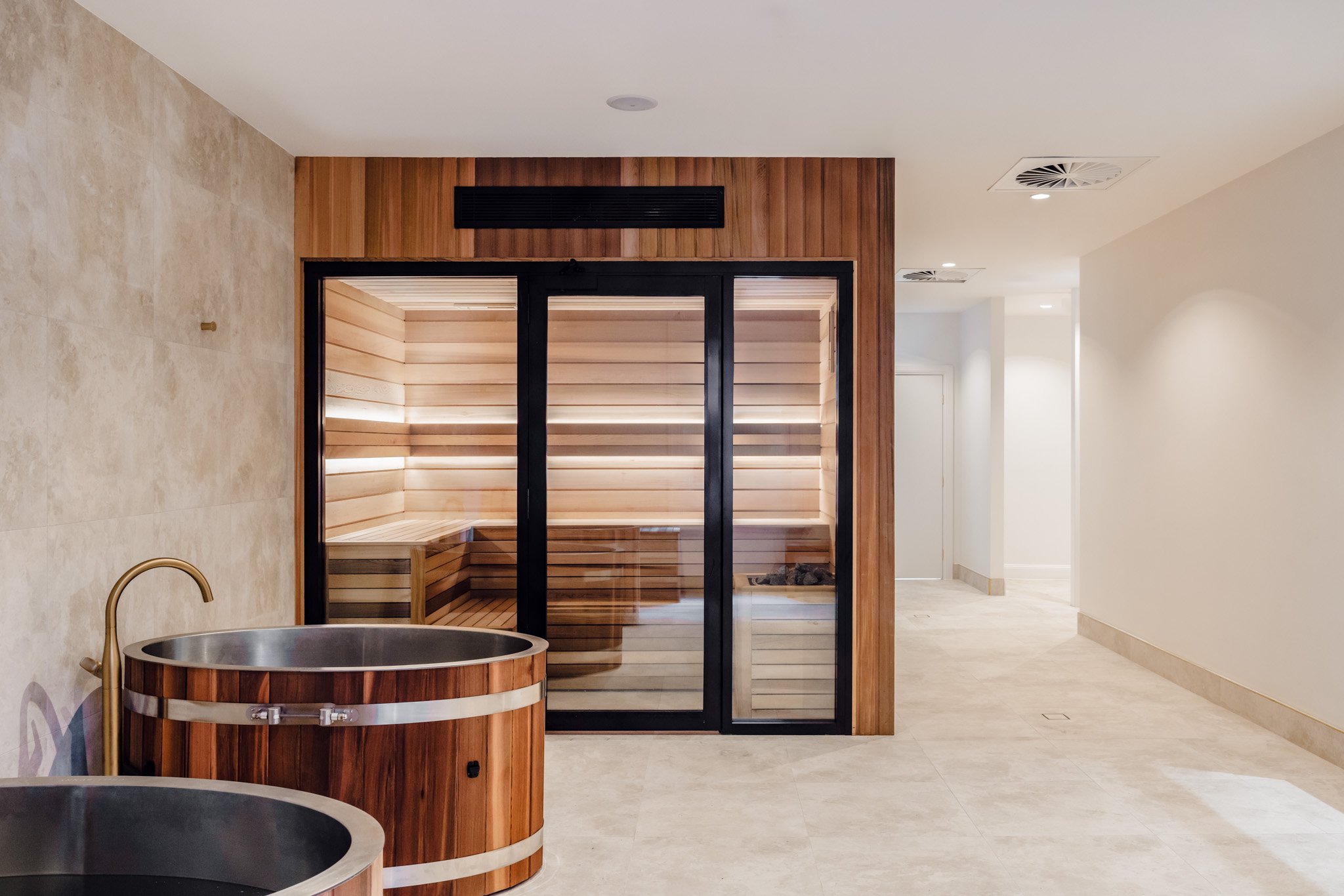 Modern indoor sauna room with glass doors and wooden interior; adjacent lounging tubs with wood surrounds and gold fixtures in a neutral-colored spa area.