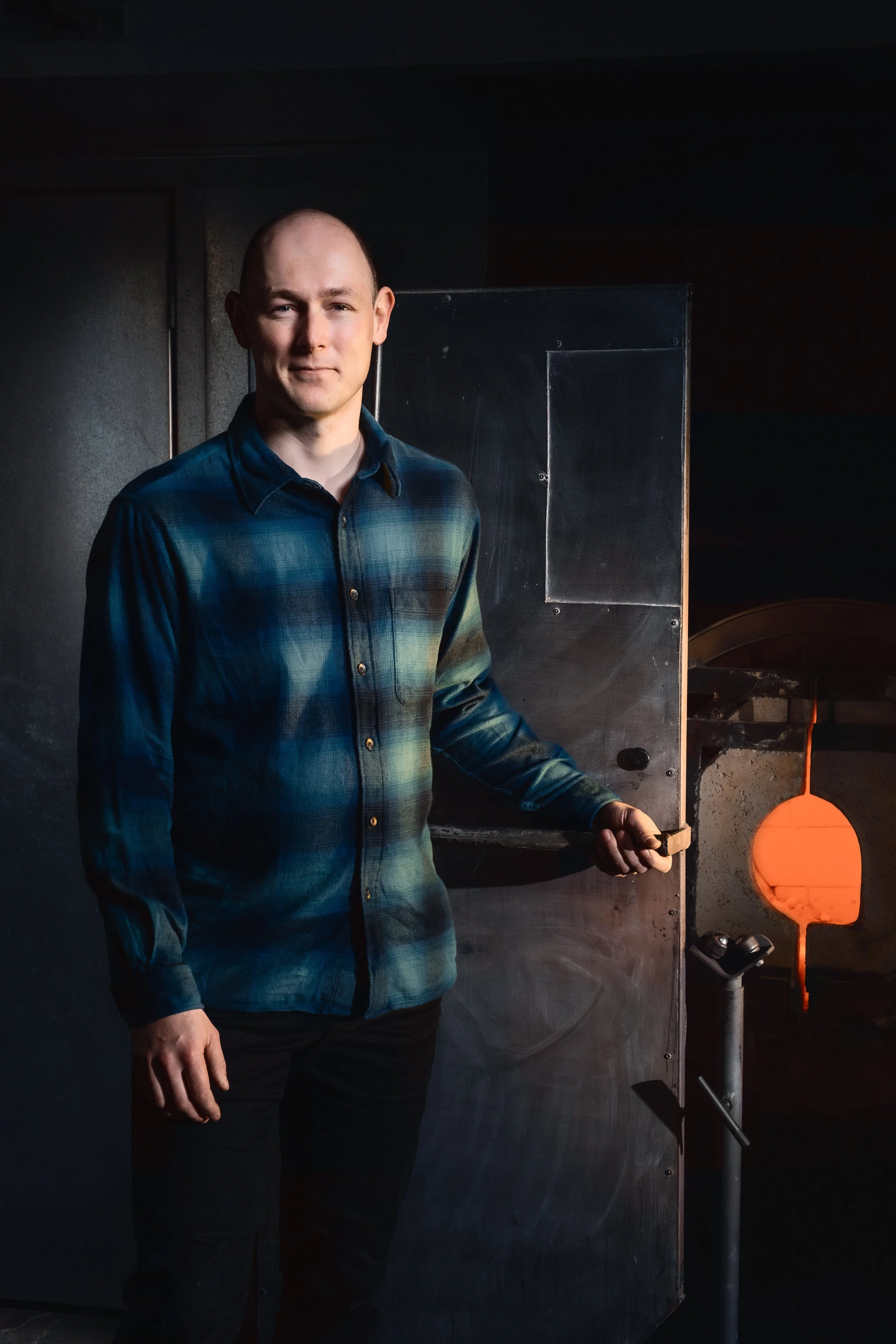 A man in a plaid shirt standing in front of a furnace with a glowing interior, holding a tool.