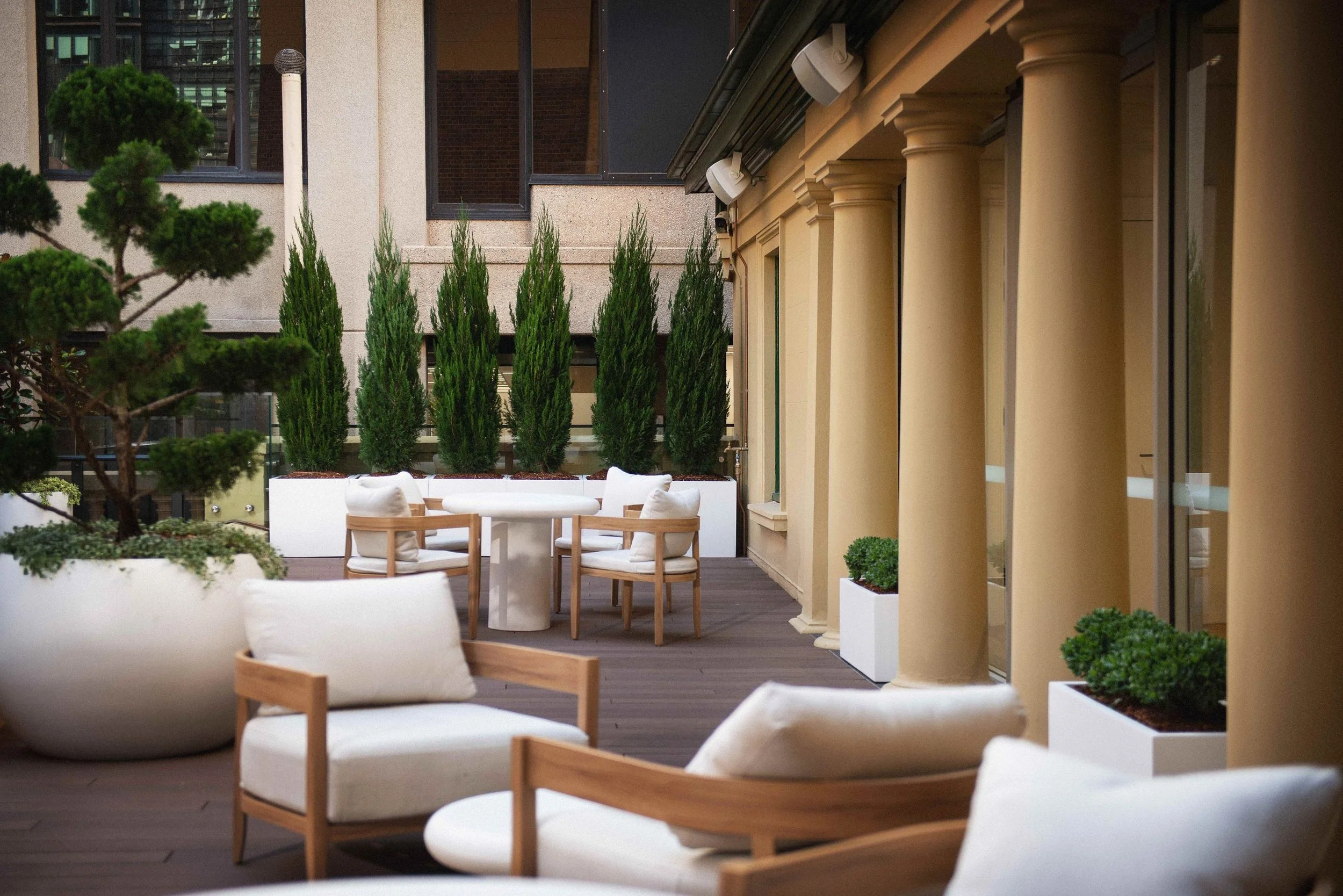 A stylish outdoor terrace with cream and wooden furniture, potted plants, and tall green trees against a city building background.