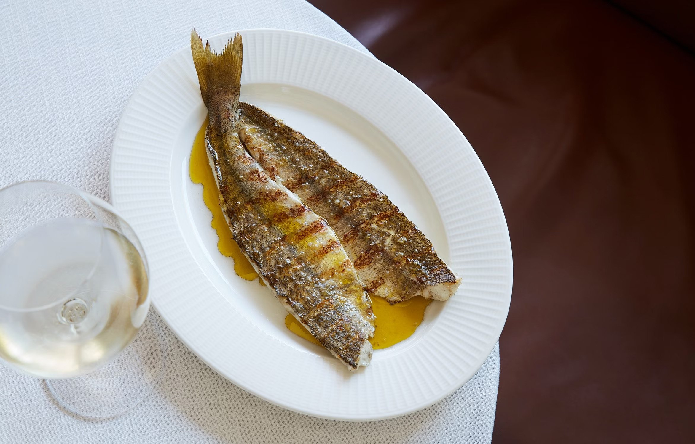 Two grilled fish fillets on a white plate with olive oil, served with a glass of white wine on a table.
