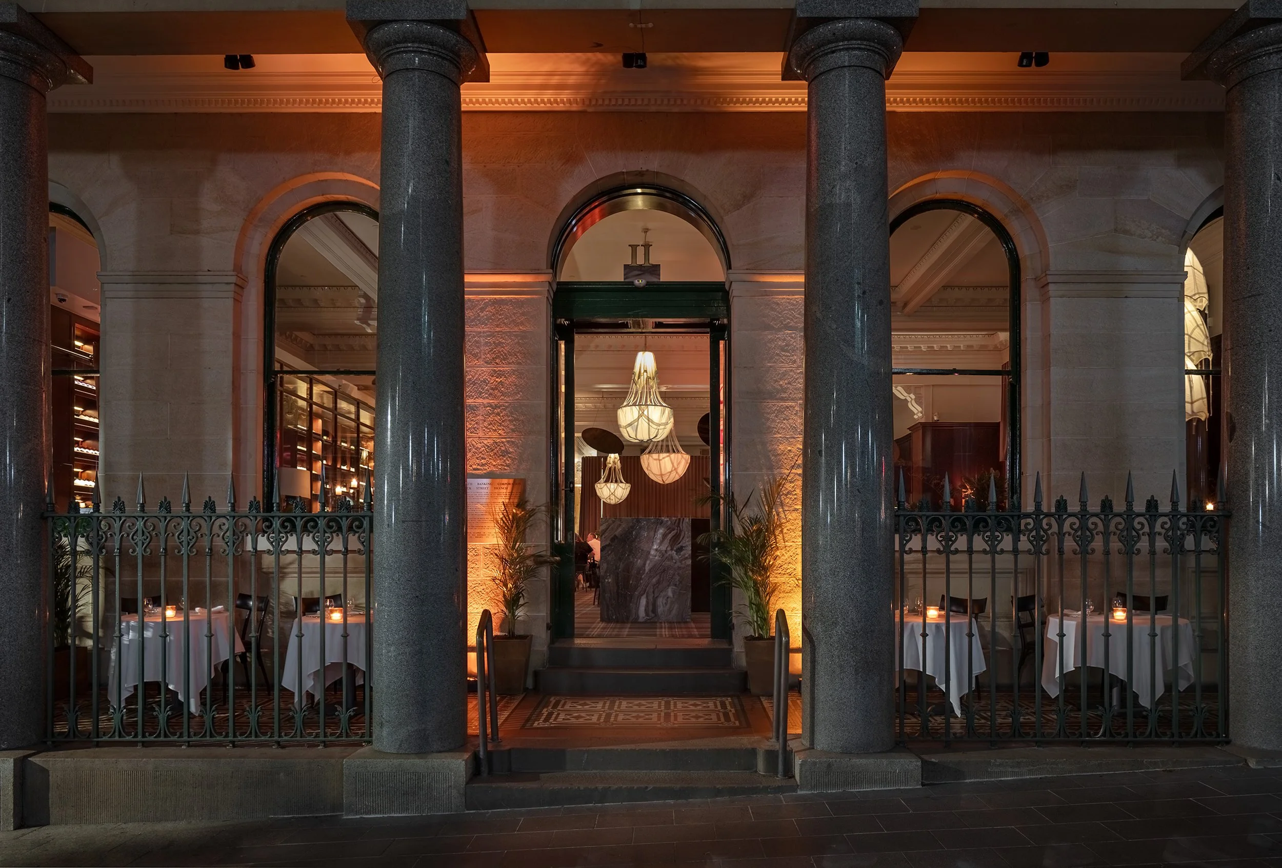 Elegant restaurant entrance with large stone columns, arched windows, and a wrought iron gate. Inside, tables are set with white tablecloths and candles, illuminated by hanging pendant lights.