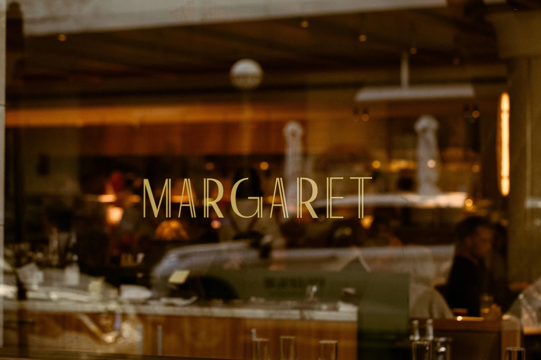 Restaurant window with the word 'MARGARET' in gold letters, showing a busy interior with people dining inside.