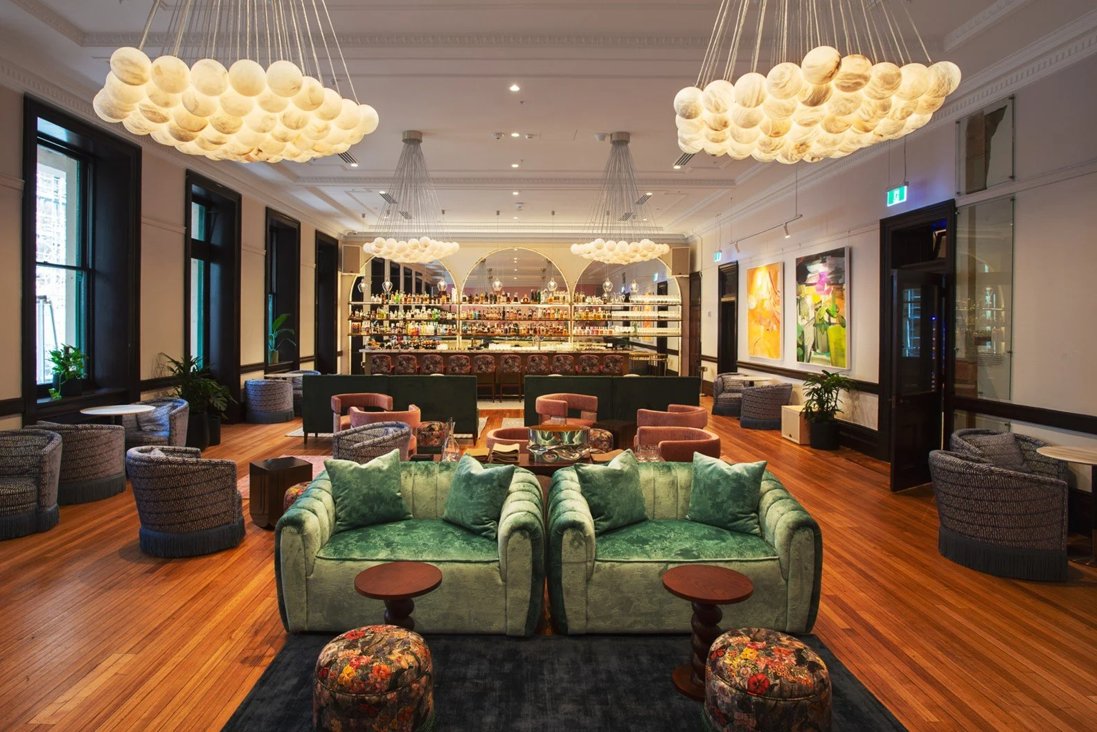 Elegant lounge with green velvet sofas, patterned armchairs, modern pendant lights resembling clouds, a well-stocked bar in the background, and colorful artwork on the walls.
