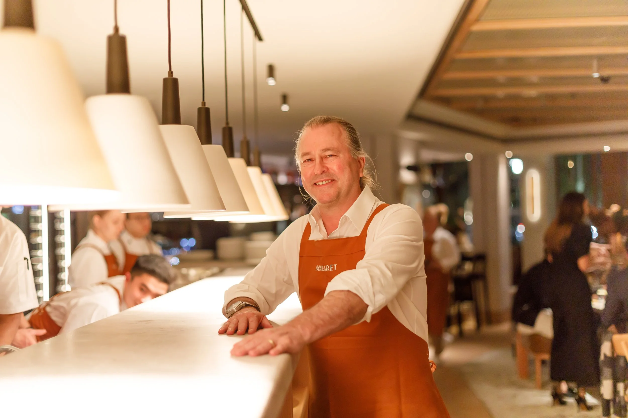 A smiling man in a white shirt and orange apron standing at a restaurant bar counter with his hands resting on it, with staff and patrons in the background.