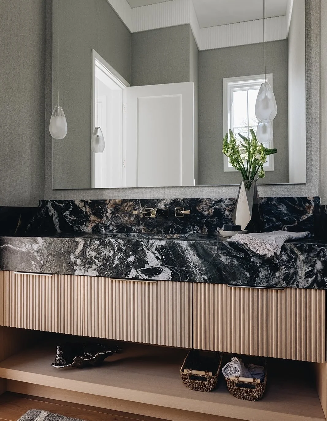 Small space, big impact.
This powder room blends bold materiality with refined detailing&mdash;from the striking stone countertop to the custom fluted cabinetry finished with sleek edge pulls. Paired pendant lights frame the vanity with understated e