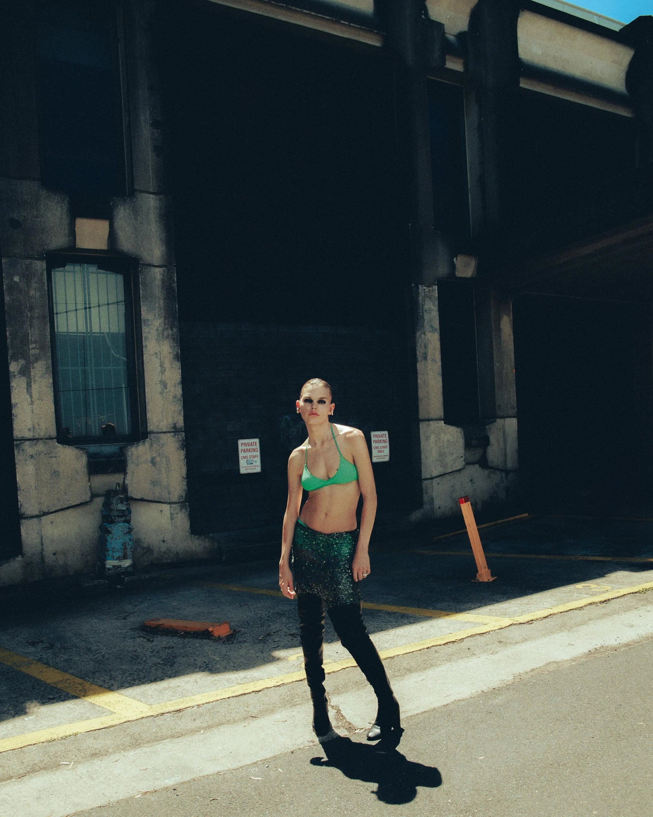 A woman standing in an urban parking lot with shadows cast on her. She is wearing a green bikini top, black shiny pants, and black shoes, with a serious expression.