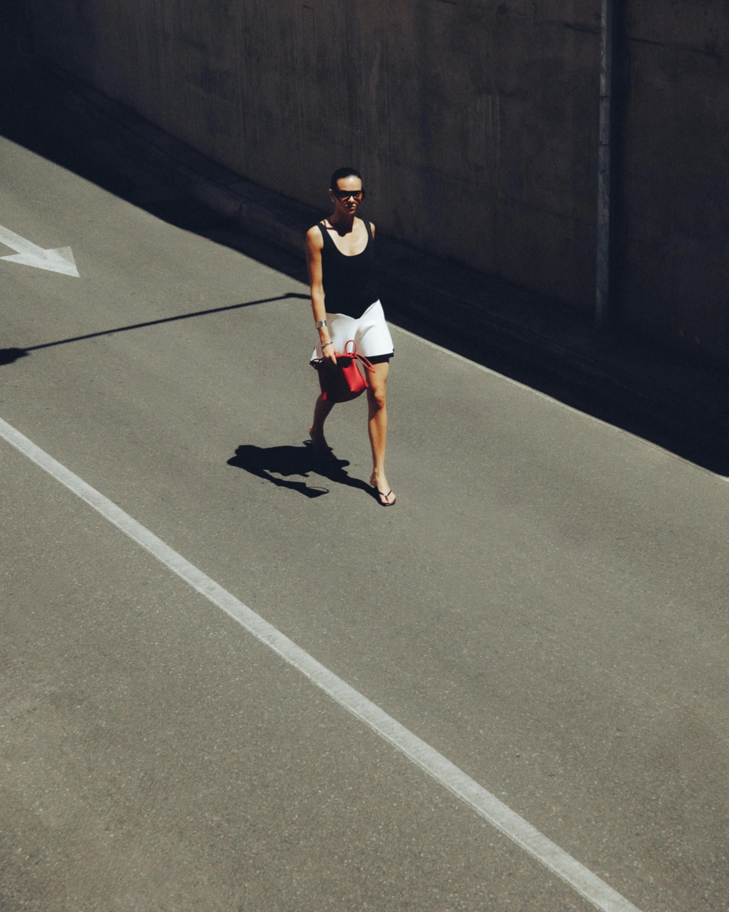 A woman walking on an empty road next to a concrete wall, carrying a red handbag, wearing sunglasses, a black sleeveless top, white skirt, and flip-flops.