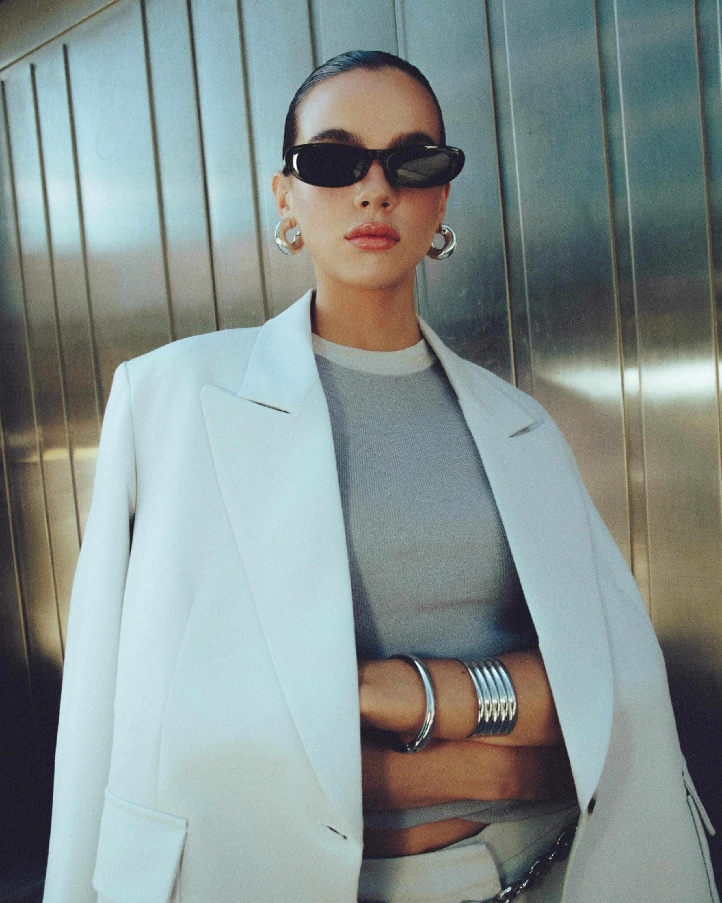 A woman with dark hair pulled back, wearing black sunglasses, hoop earrings, a light-colored blazer, a gray crop top, and multiple silver bangles on her wrist, standing in front of a metal-paneled wall.