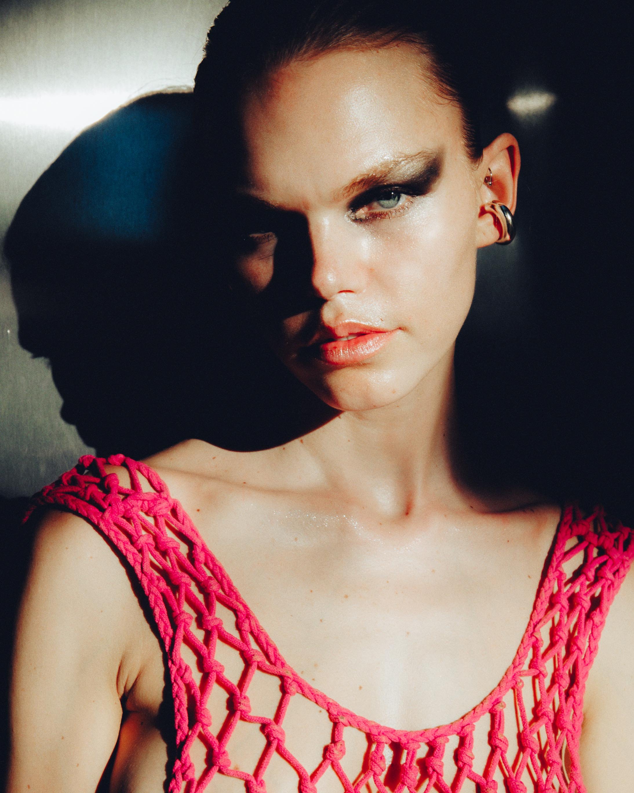 A woman with short hair and earrings wearing a pink, open-knit top, against a dark background, with dramatic lighting highlighting her face and shoulder.