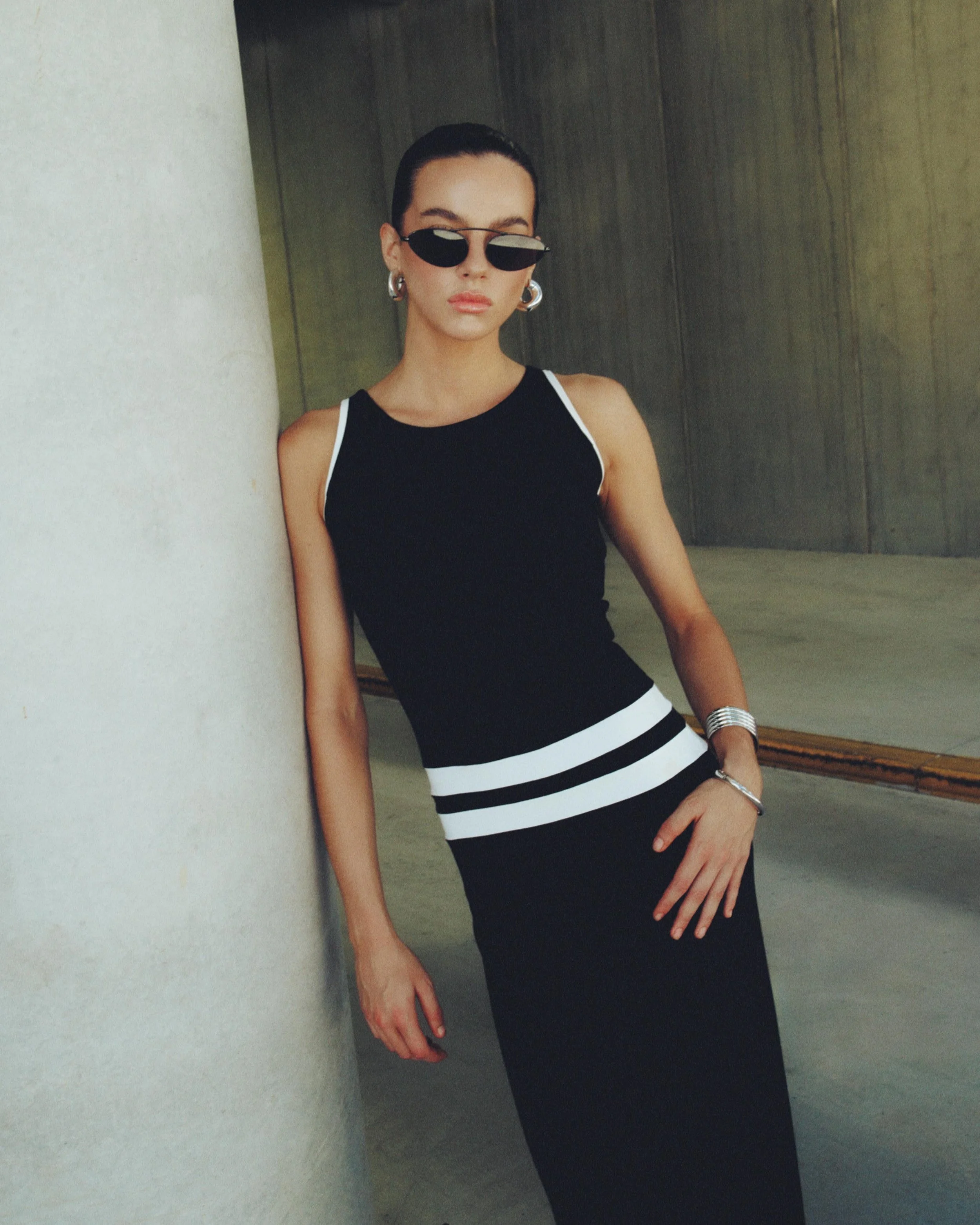 A woman wearing a black sleeveless top with white trim and a matching black skirt with white stripes at the waist, leaning against a white pillar indoors, accessorized with large hoop earrings, sunglasses, and a watch.