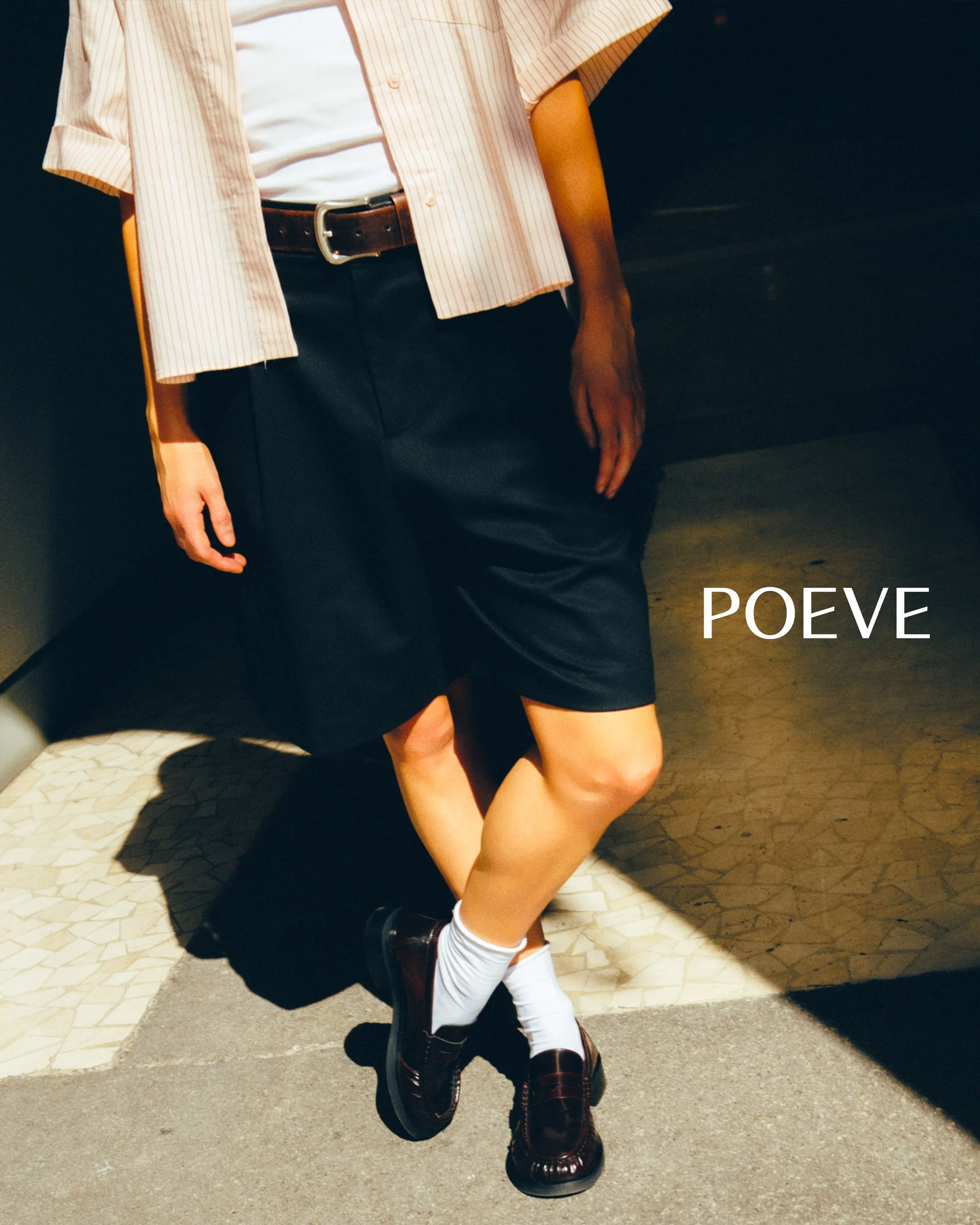 Person wearing a beige striped shirt, black shorts, white socks, and dark loafers, standing next to a couch with sunlight casting a shadow, with the word "POEVE" written on the image.