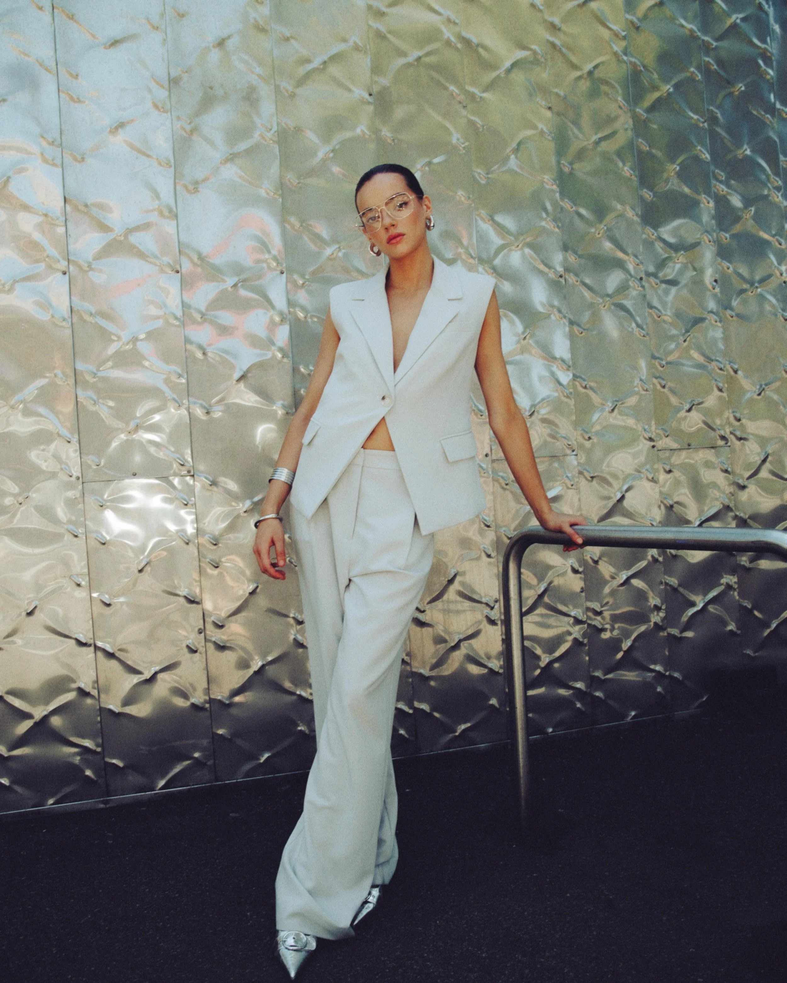 A woman in a white sleeveless blazer and wide-leg pants leaning against a metal railing in front of a metallic wall.