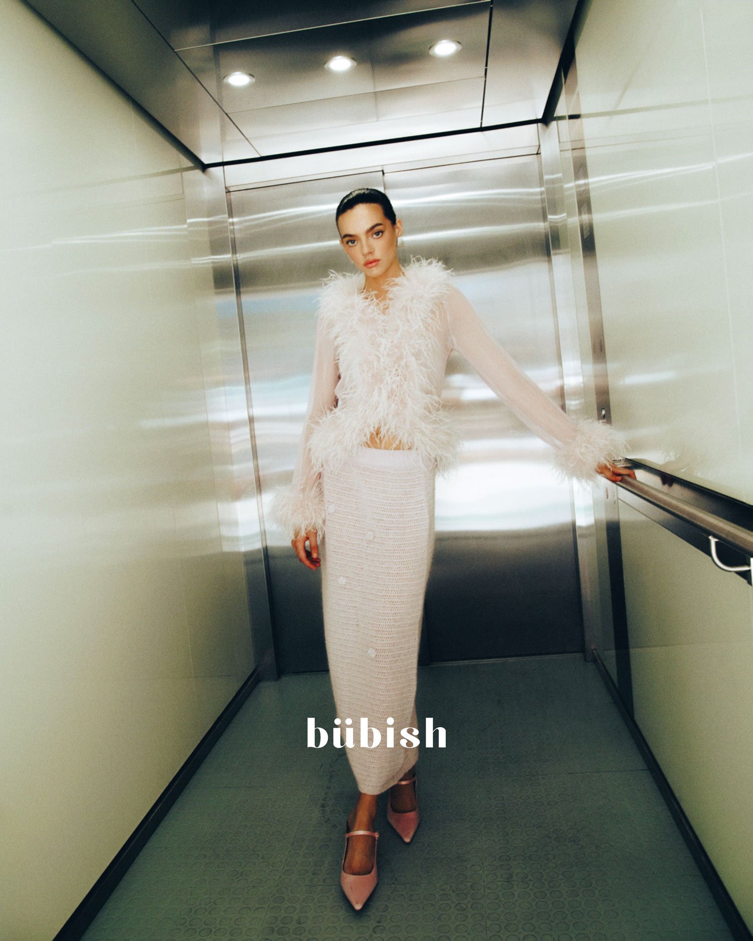 A woman with dark hair pushed back, dressed in a light pink feathered top and a white textured skirt, standing in an elevator with reflective metal walls, holding the handrail and looking at the camera.