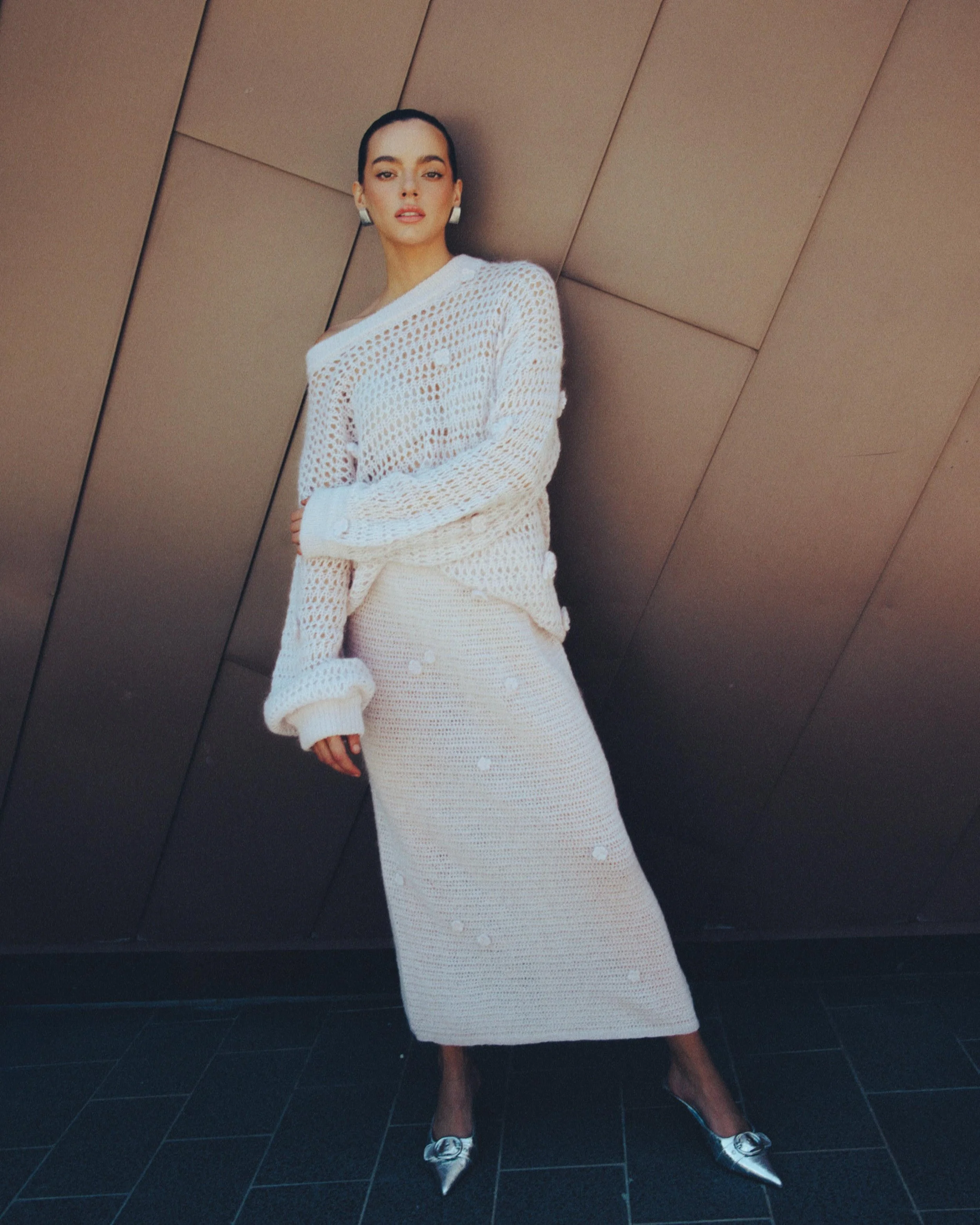 Woman wearing a white crochet sweater and skirt, standing against a brown panel wall.