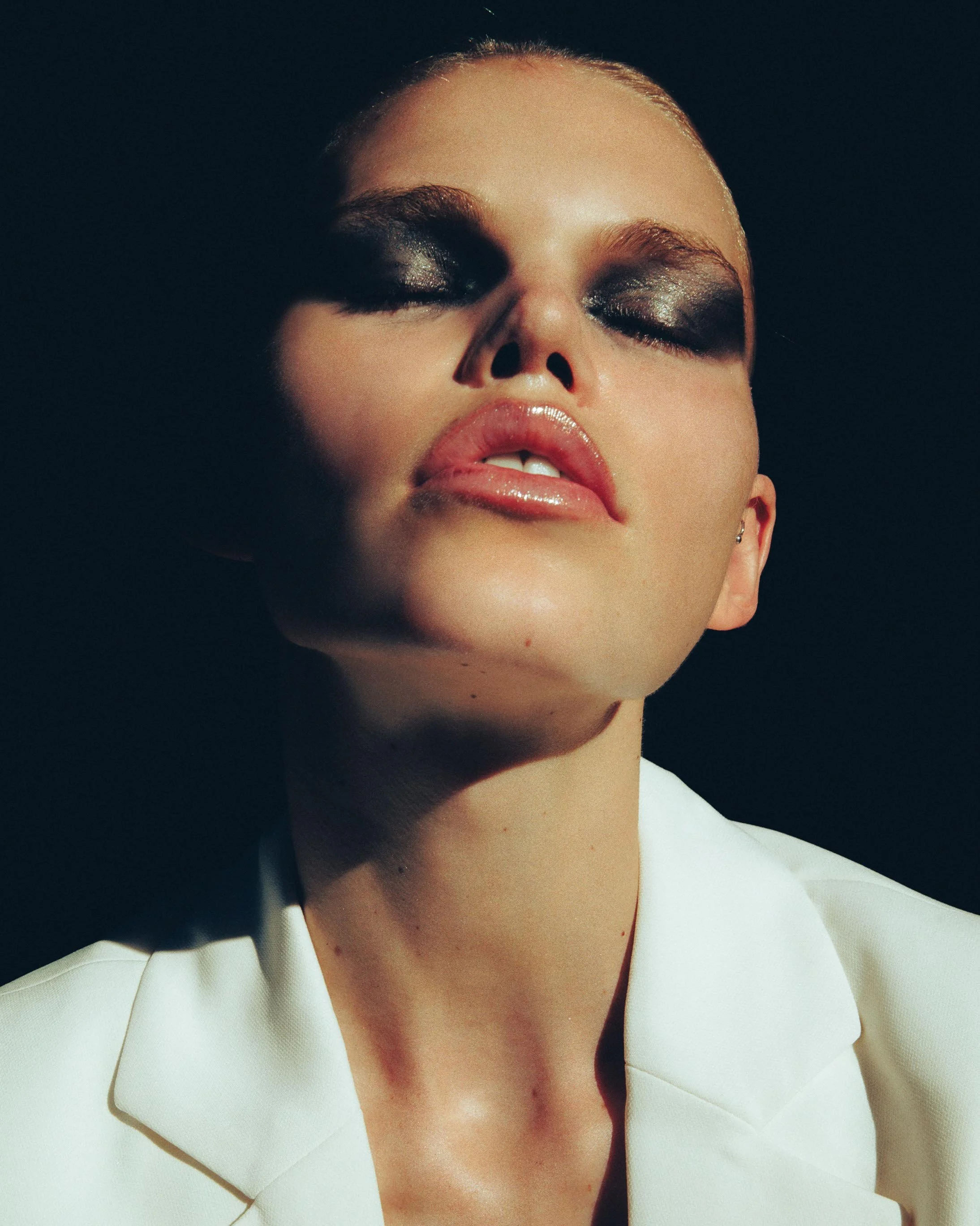 Close-up of a woman with dark makeup, glossy lips, and wearing a white blazer against a dark background.
