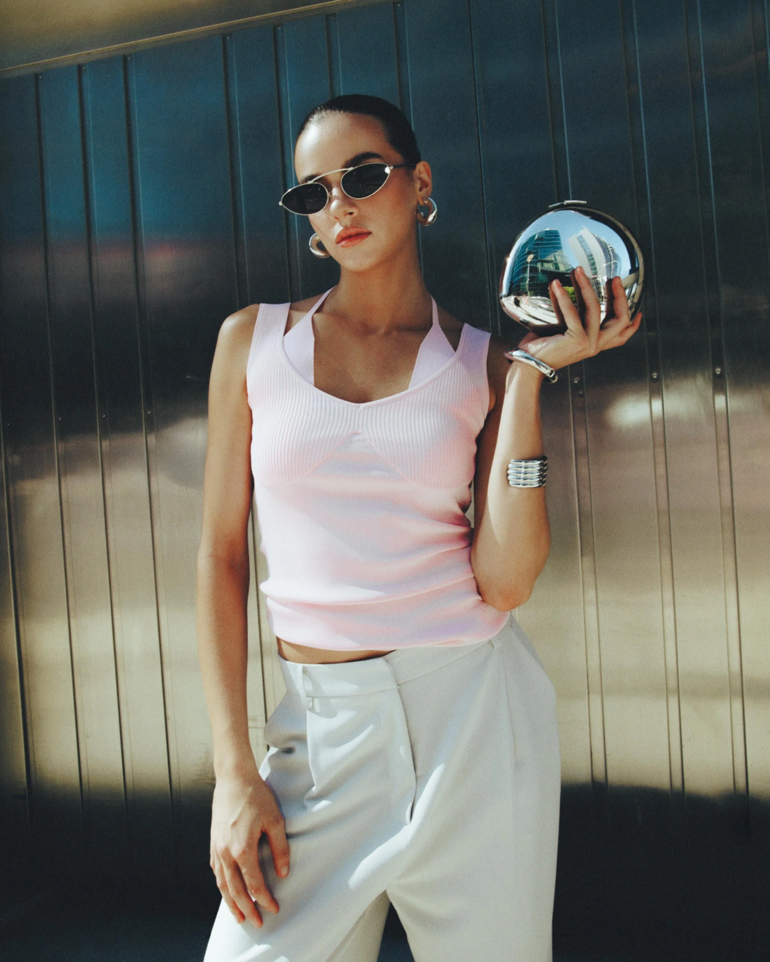 A woman in sunglasses, a pink sleeveless top, and white pants standing outdoors against a steel wall, holding a shiny, reflective metallic sphere in her right hand.
