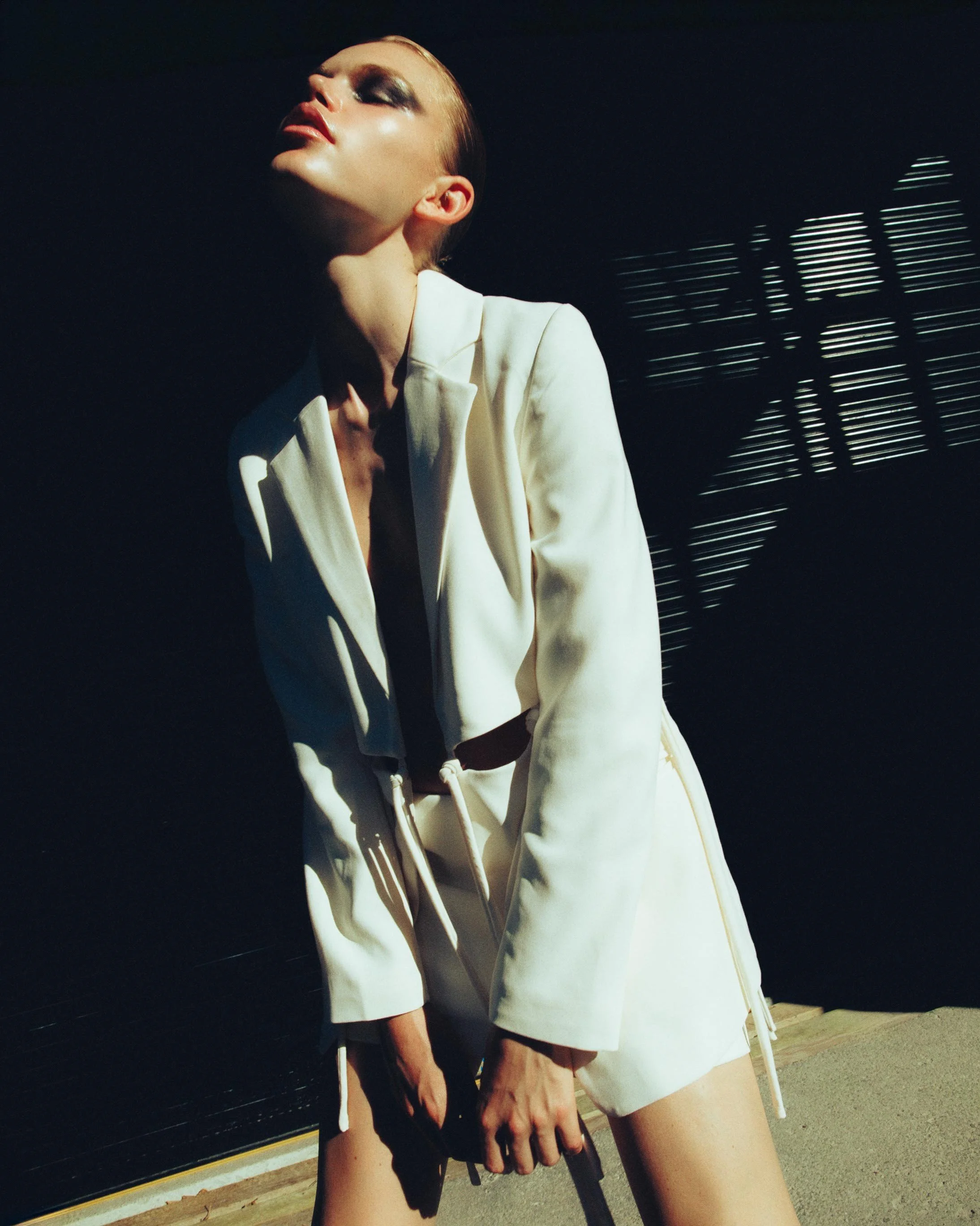 A woman wearing a white blazer and shorts, posing with her head tilted back and eyes closed, with sunlight casting shadows on her face and a blurred background.