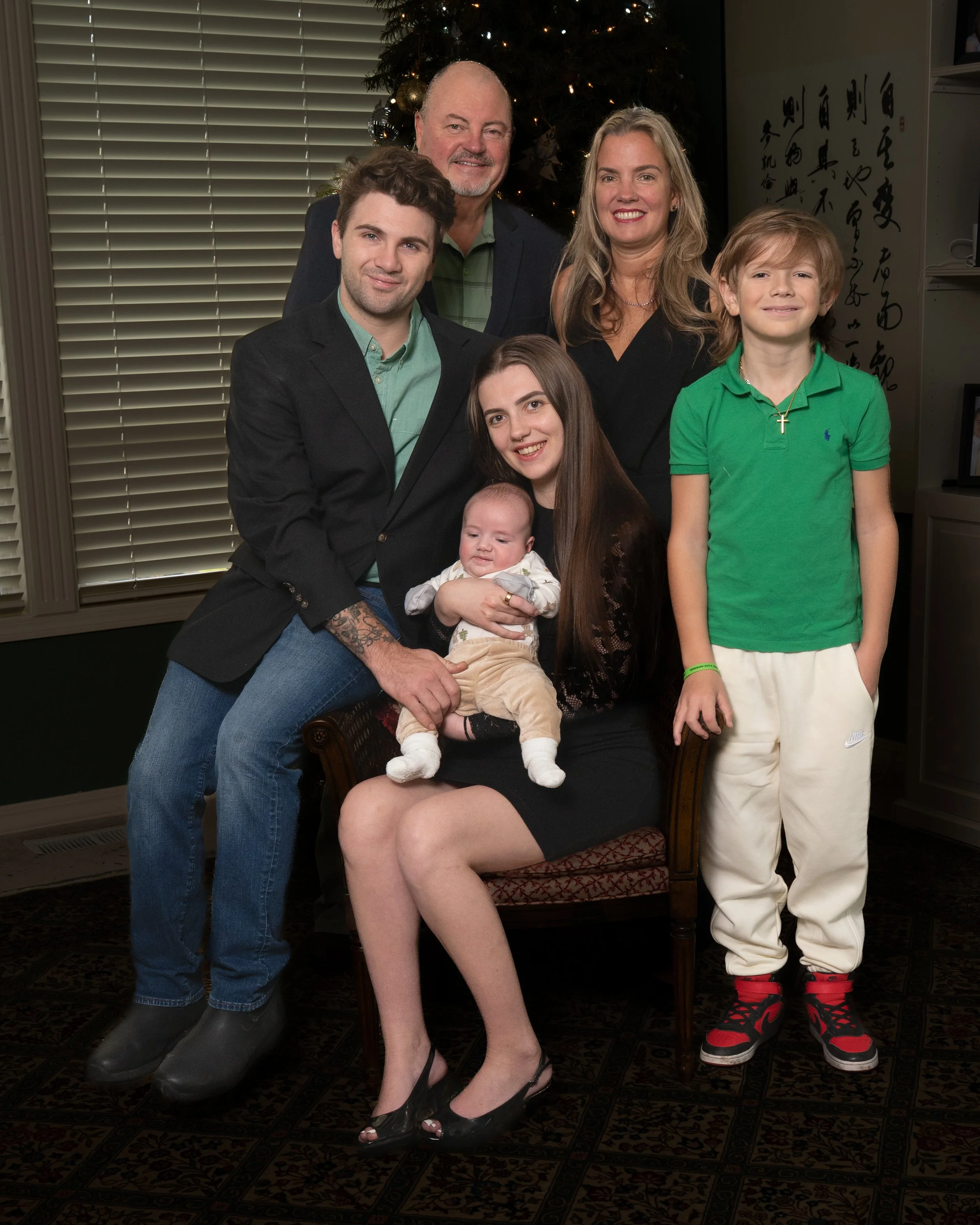 A family portrait of seven people, posing indoors in front of a Christmas tree, with a window with blinds behind them.