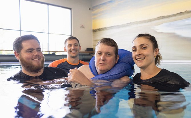 Male I Am Here resident in a swimming pool hydrotherapy session, with smiling team