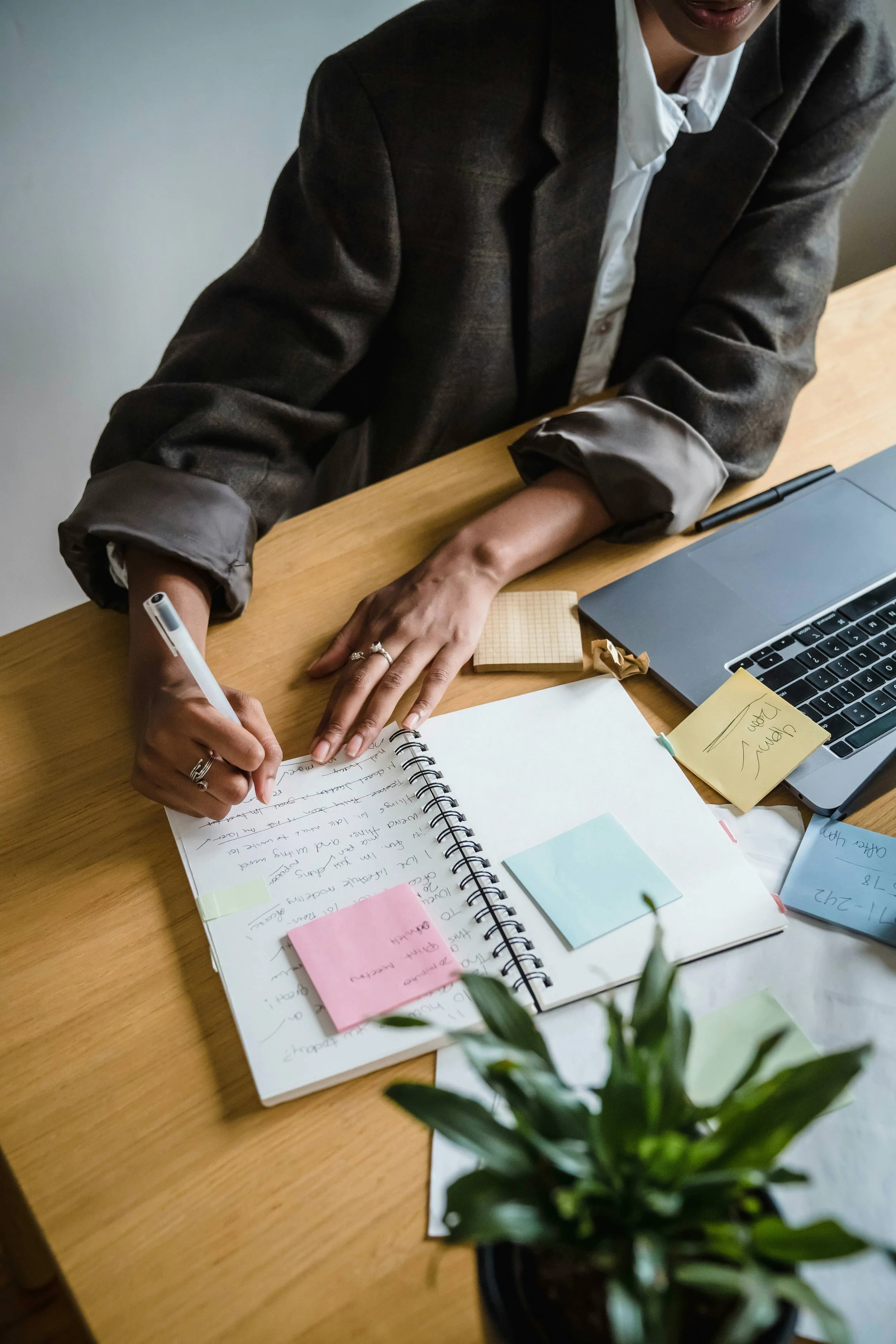 A person at a wooden desk writing in a spiral notebook with several sticky notes, surrounded by a laptop, notepad, pen, and a potted plant in the foreground.