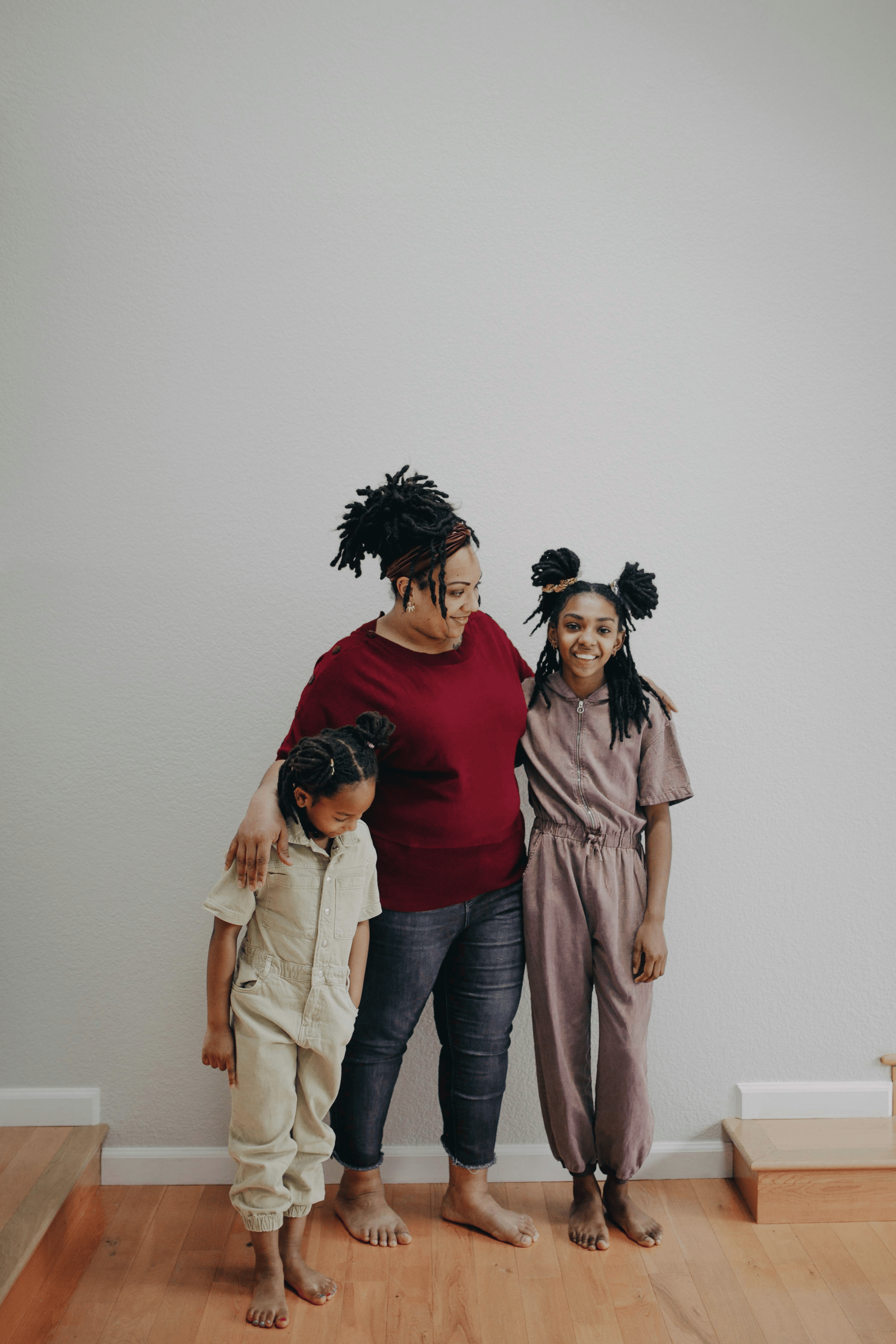 A woman with dreadlocks and wearing a red top stands with three children, holding one child's hand, in a room with a white wall and hardwood floor.