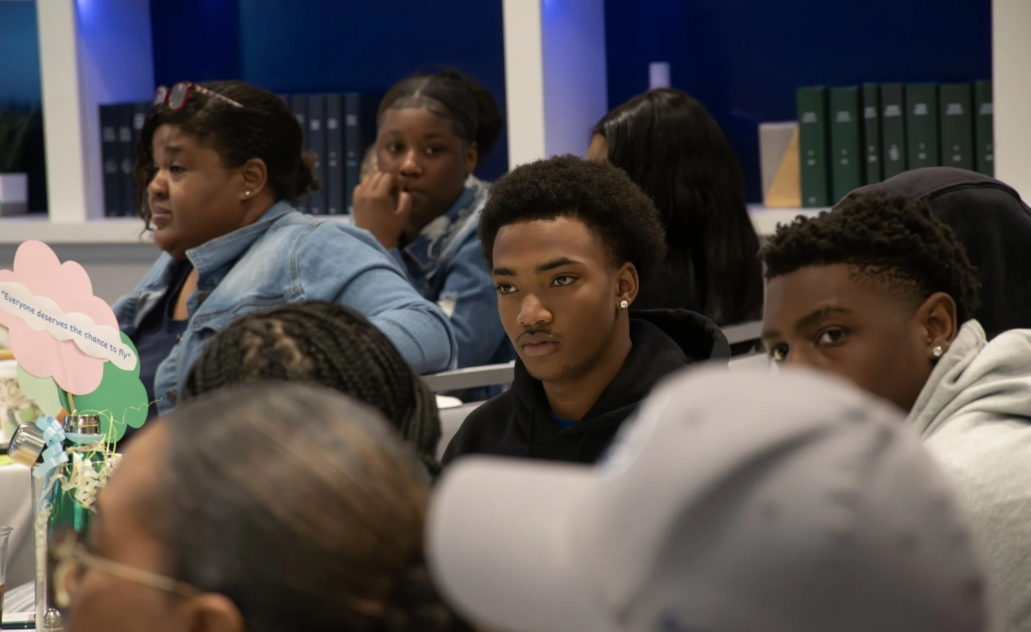 A group of young people sitting at desks, attentively listening during a classroom session, with bookshelves in the background.