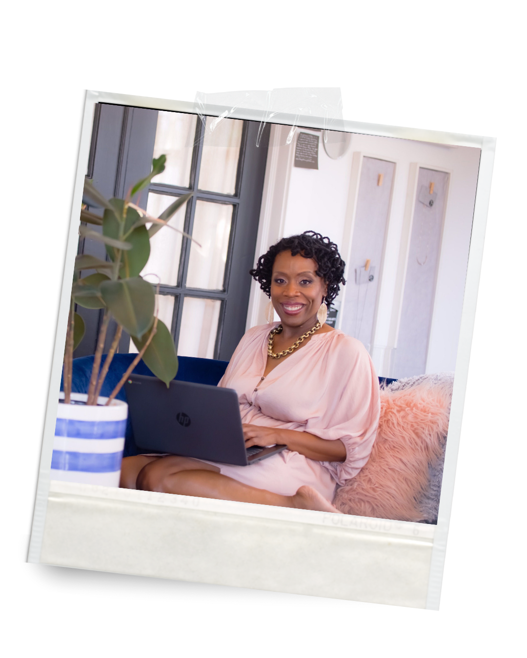 A woman, Gretchen Campbell, sitting on a cozy pink fur blanket, holding a laptop, smiling, with a potted plant in the foreground, in a well-lit room with large windows and decorative wall panels.