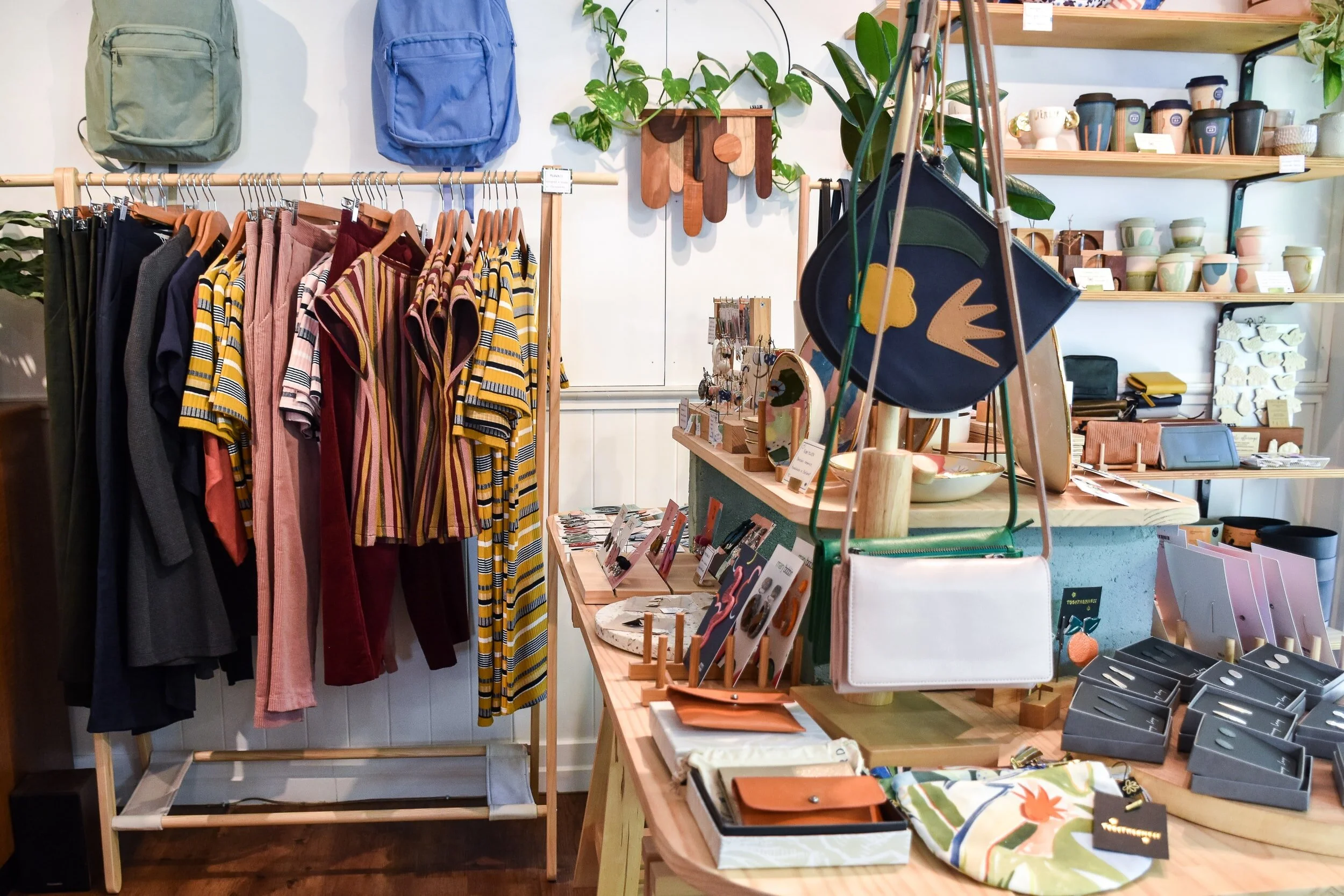 Clothing and accessories display in boutique store, featuring hanging striped shirts and pants, handbags, jewelry, ceramic cups, and decorative items on shelves.