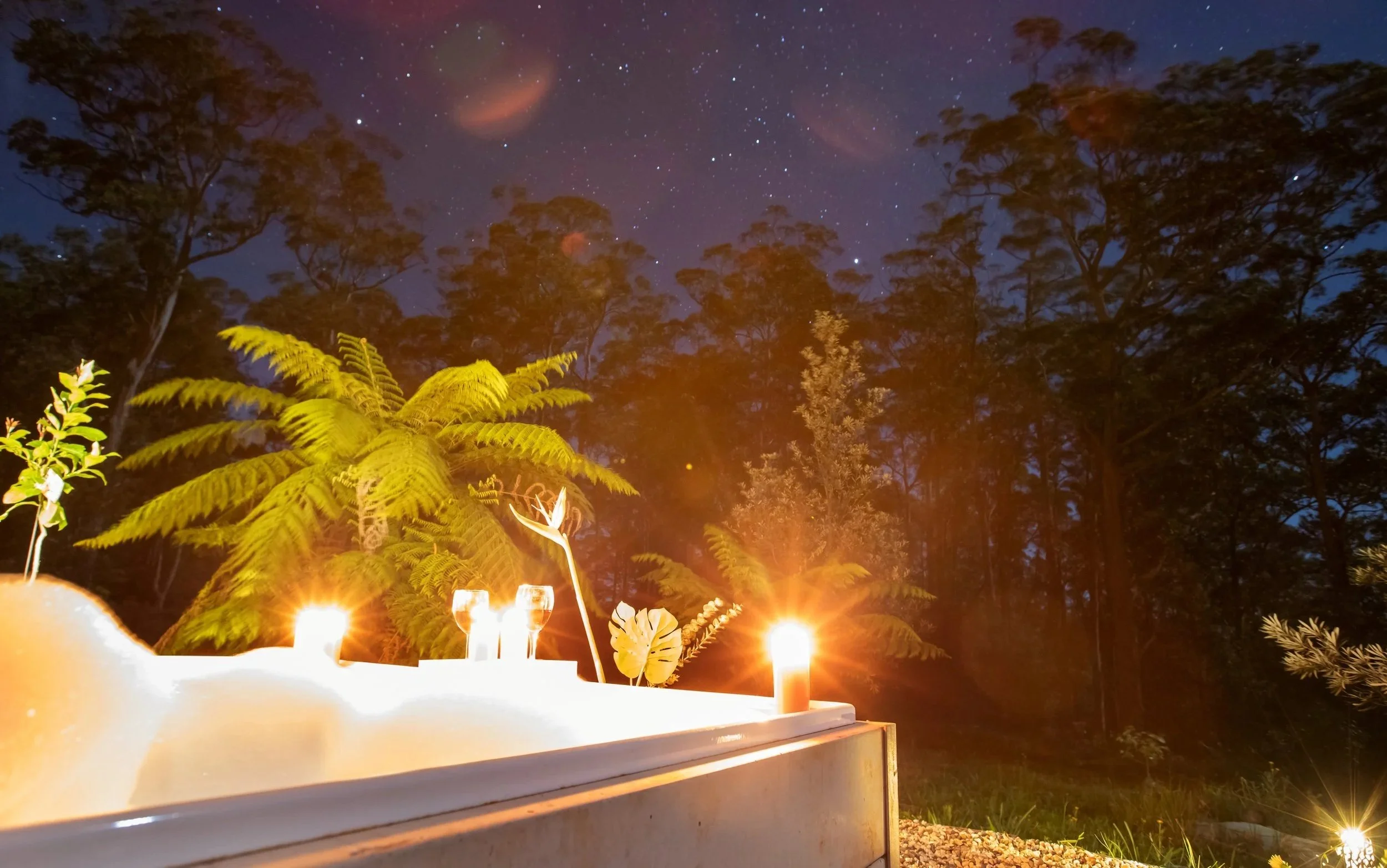 Outdoor night scene with candles on white table, lush greenery including ferns, and tall trees under a starry sky.
