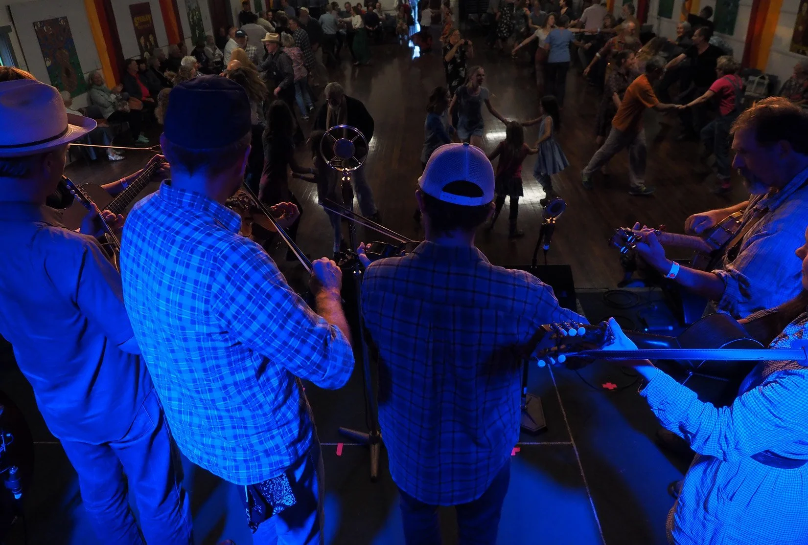 A band of four musicians playing guitars on stage, viewed from behind, with a dance floor of people dancing and socializing in the background.