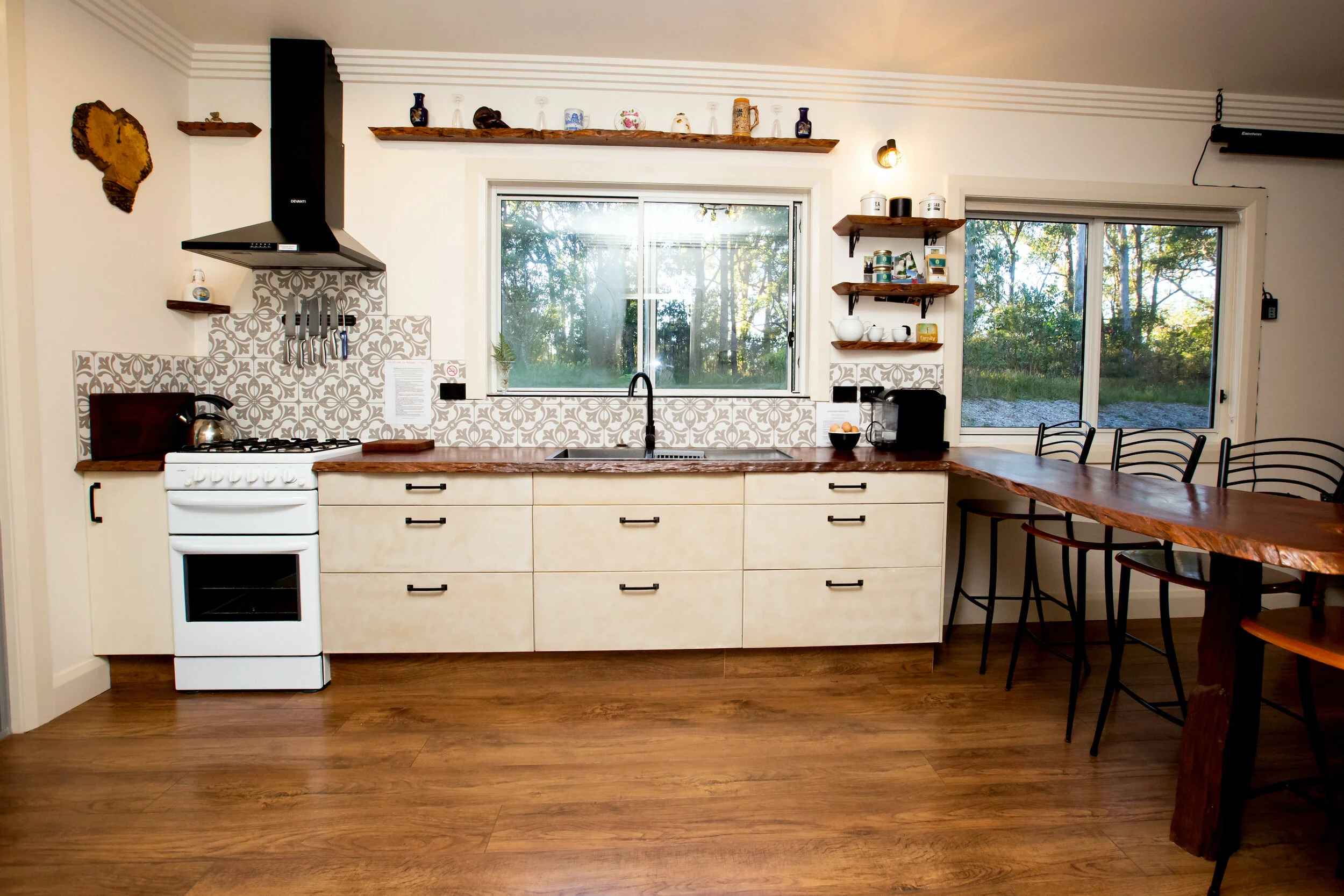 A kitchen with a white stove, a black range hood, and beige cabinets. There are two windows above the counter, one with a view of trees outside. Open wooden shelves hold jars and decorative items. A wooden dining table with black chairs is on the right. The kitchen has a wood floor and decorative tile backsplash.