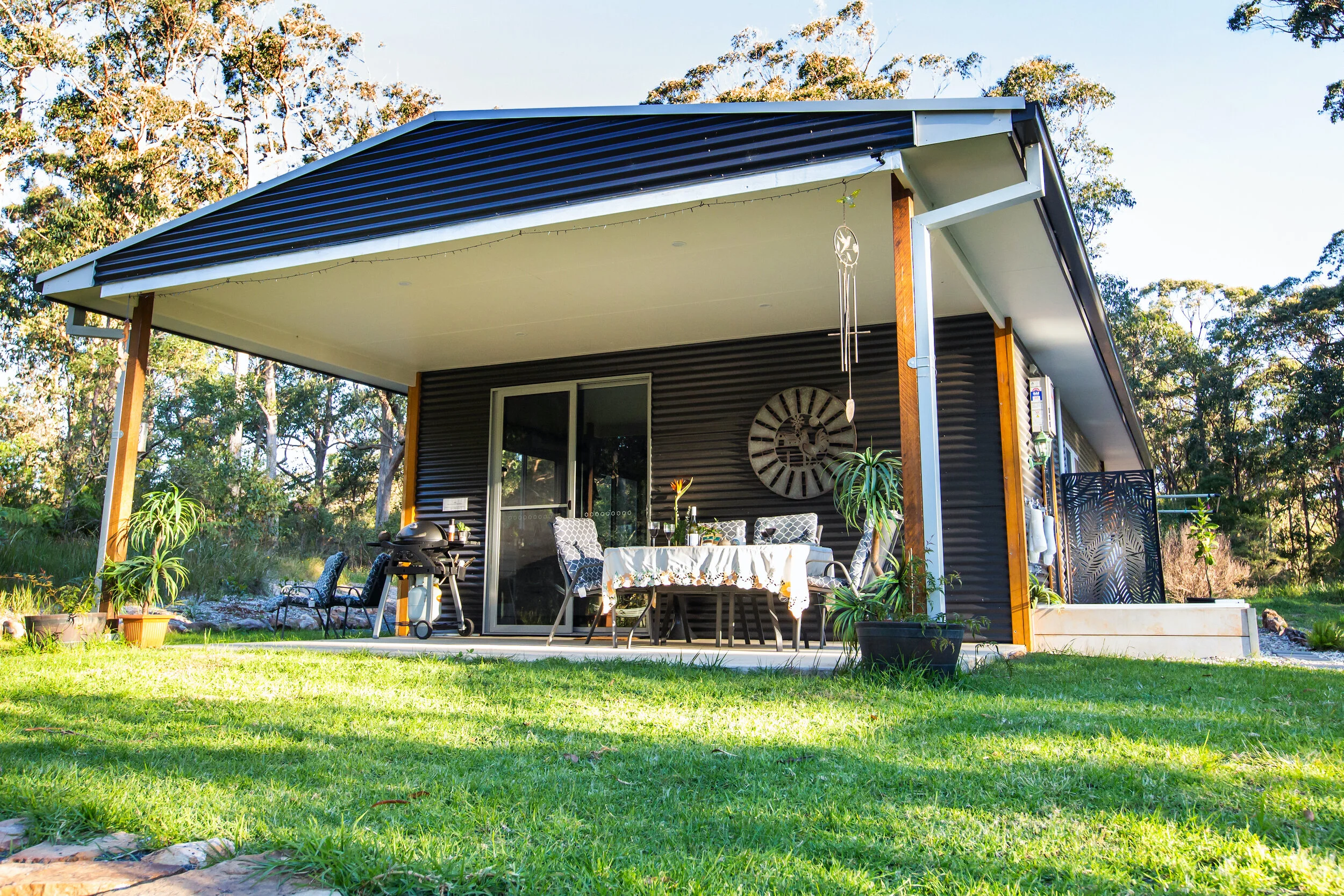 A modern house with a covered patio, outdoor dining table, and potted plants, surrounded by trees.