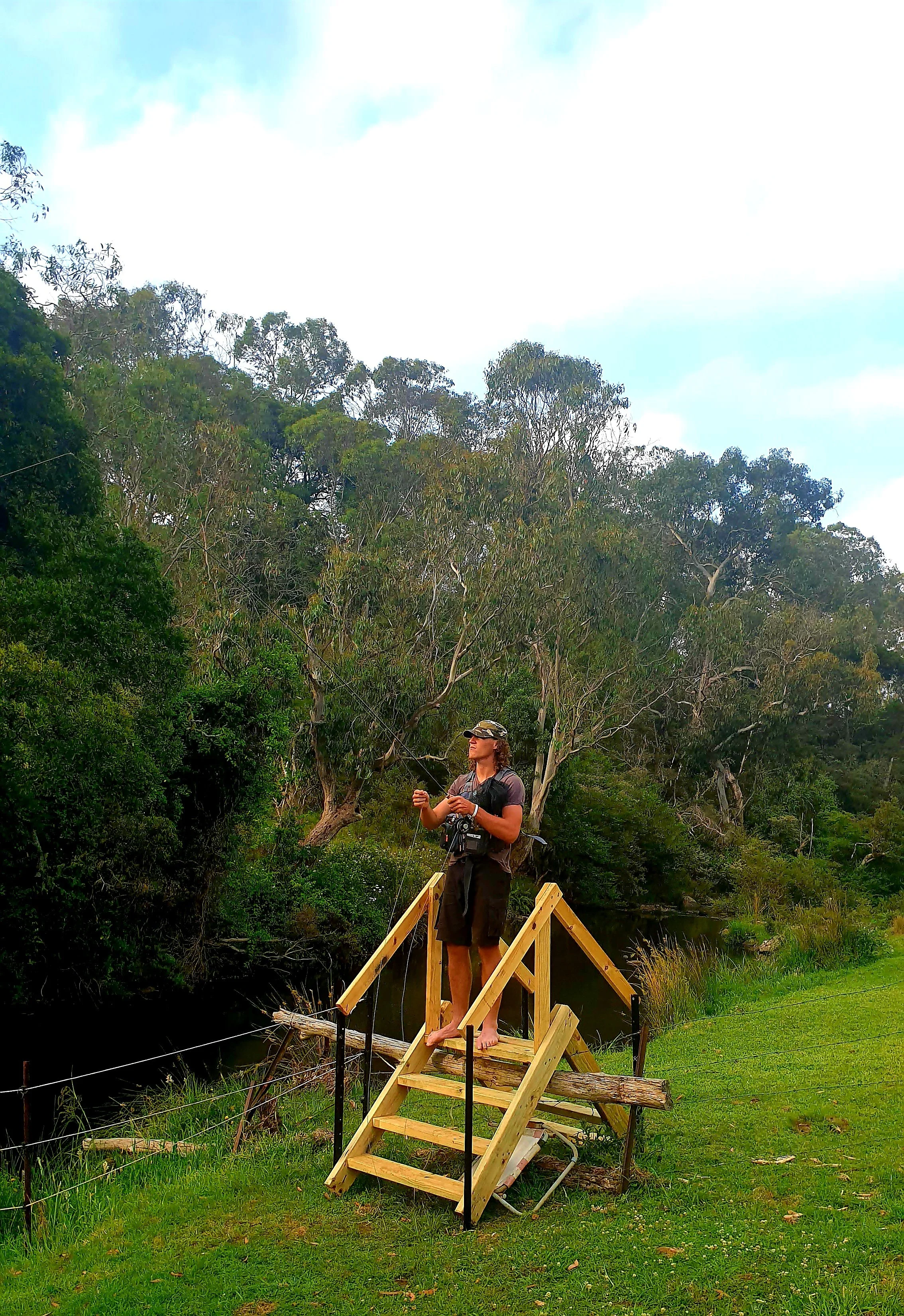 A person standing barefoot on a small wooden platform, holding a fishing rod, surrounded by lush greenery and trees, with a bright sky above.
